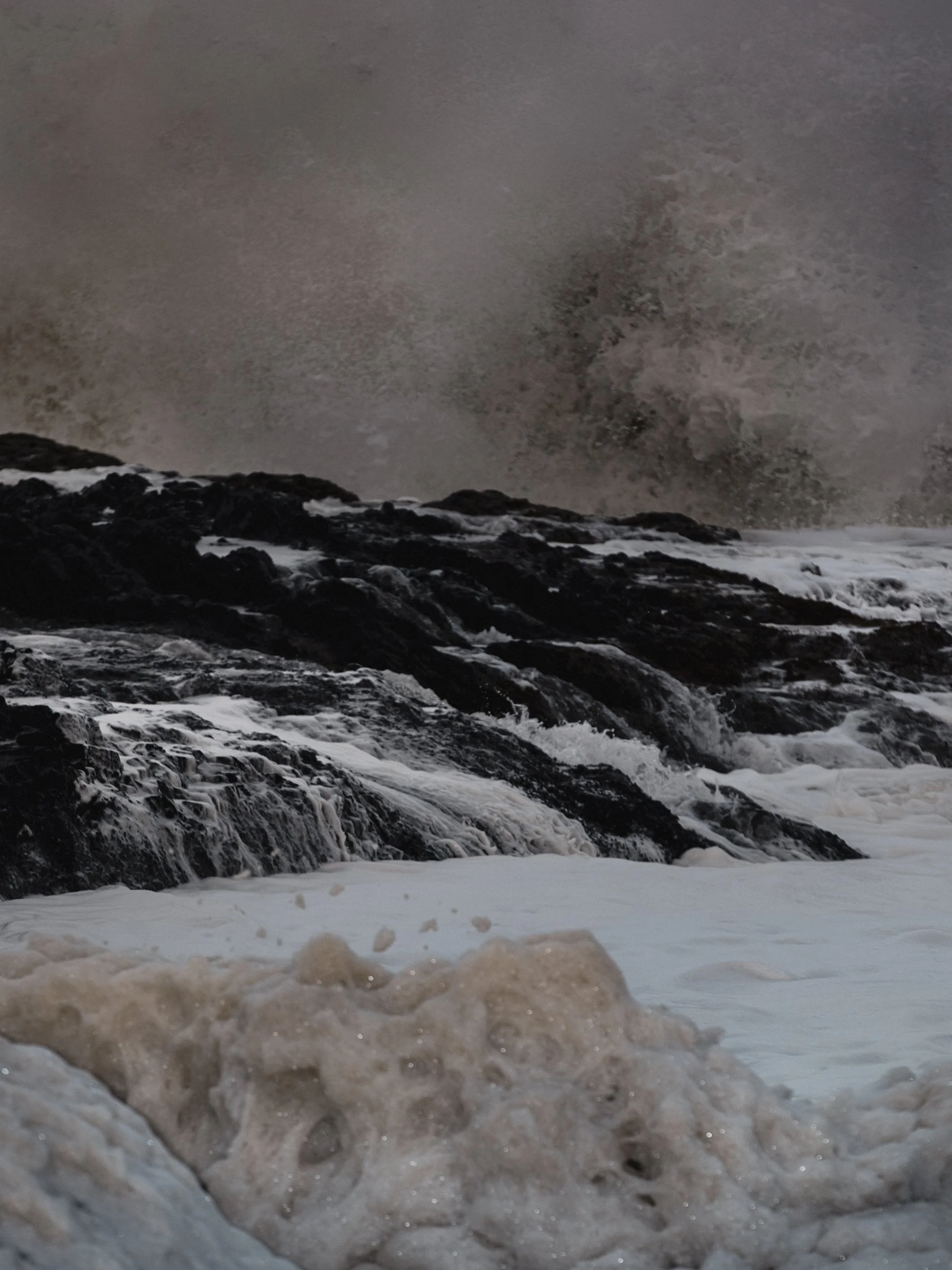 A rocky shoreline with waves crashing against dark rocks under a smoky, overcast sky.