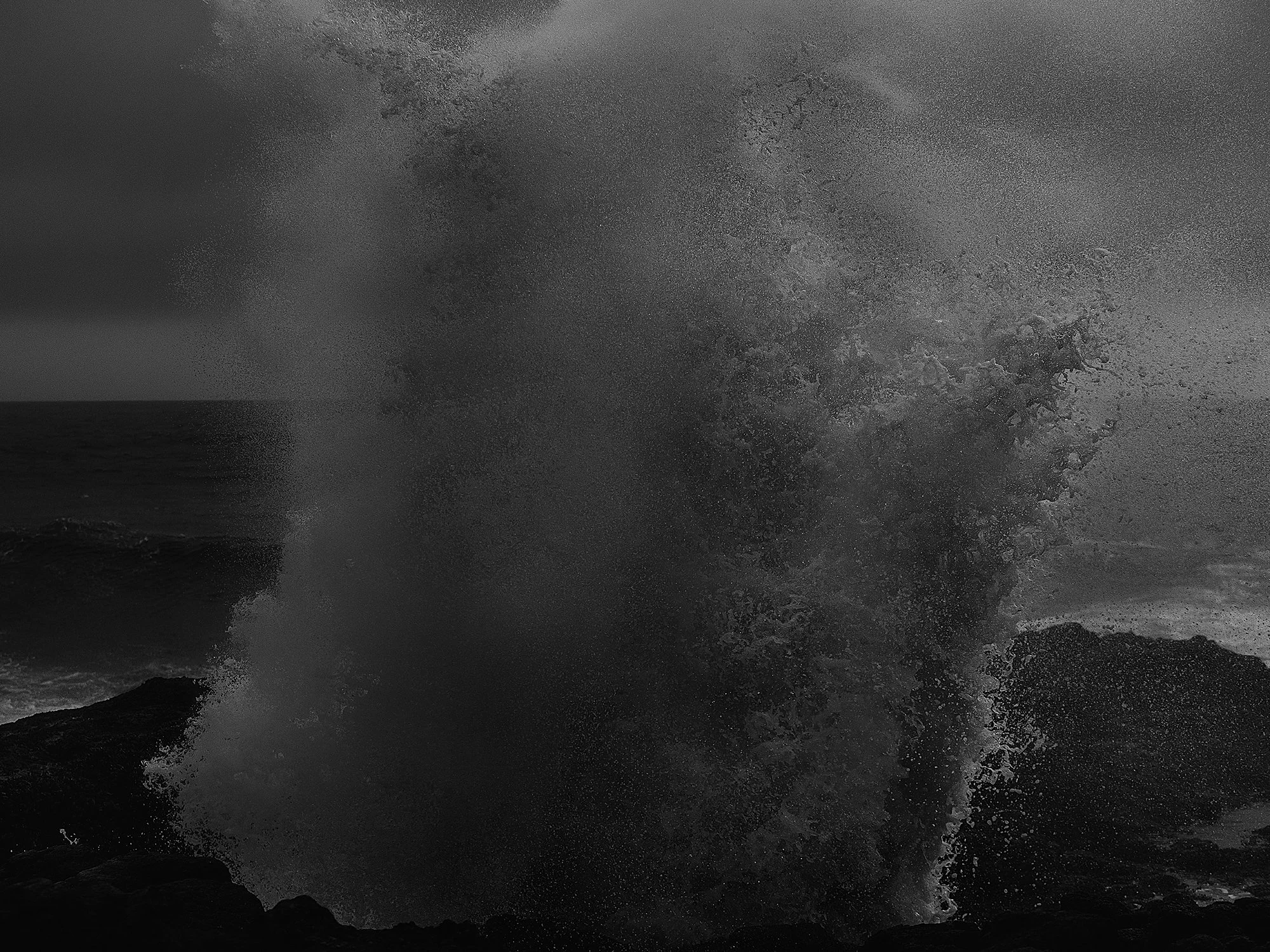 Black and white photograph of ocean waves crashing against rocks, with spray and foam in the air.