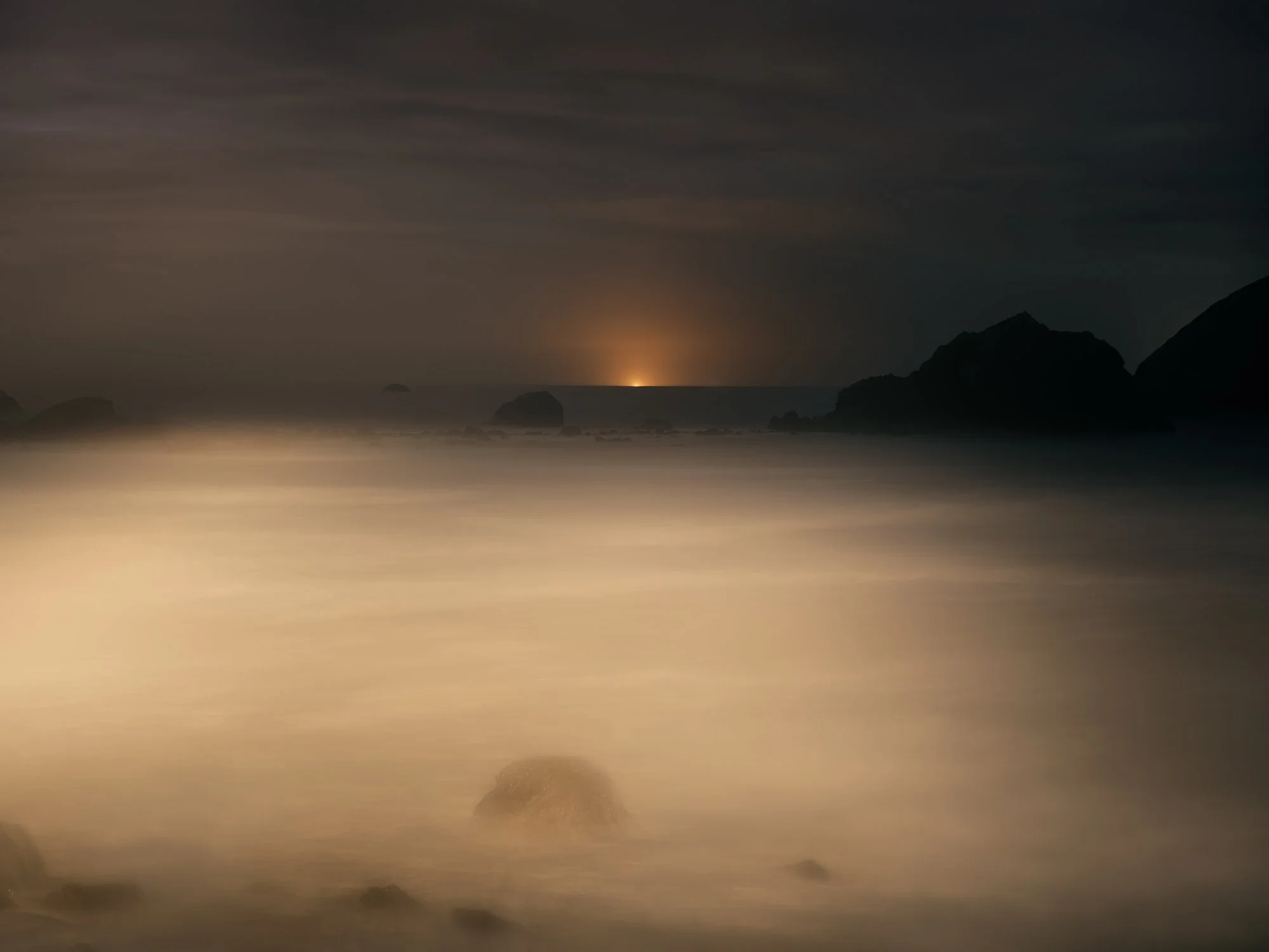 A serene coastal scene at twilight with large rocks near the shoreline, calm water, a dark mountain silhouette on the right, and a faint glow on the horizon suggesting sunset or sunrise, with the sky partly cloudy.