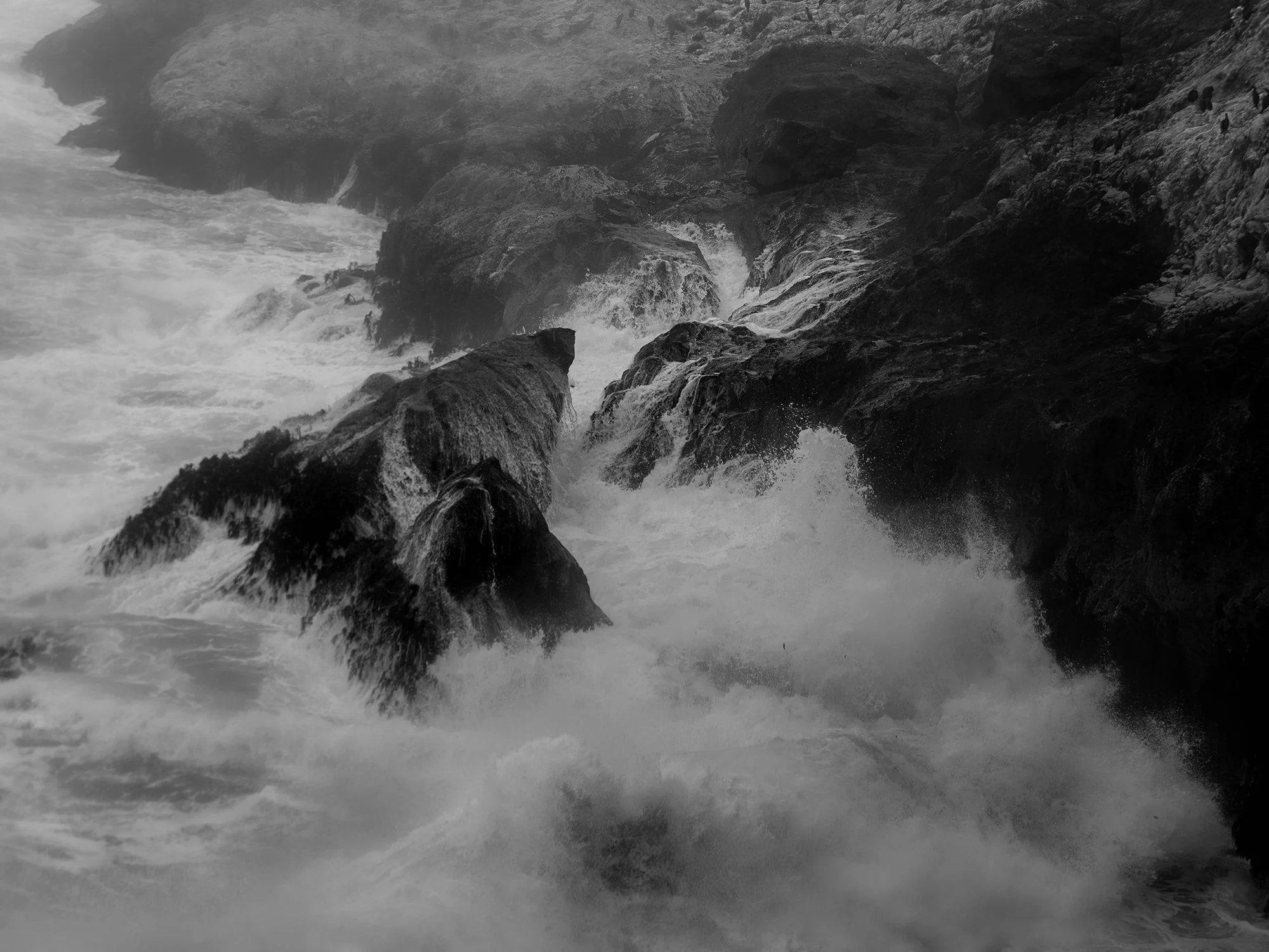 Black and white photograph of ocean waves crashing against large rocks on a rugged coastline, with waves foaming around the rocks.