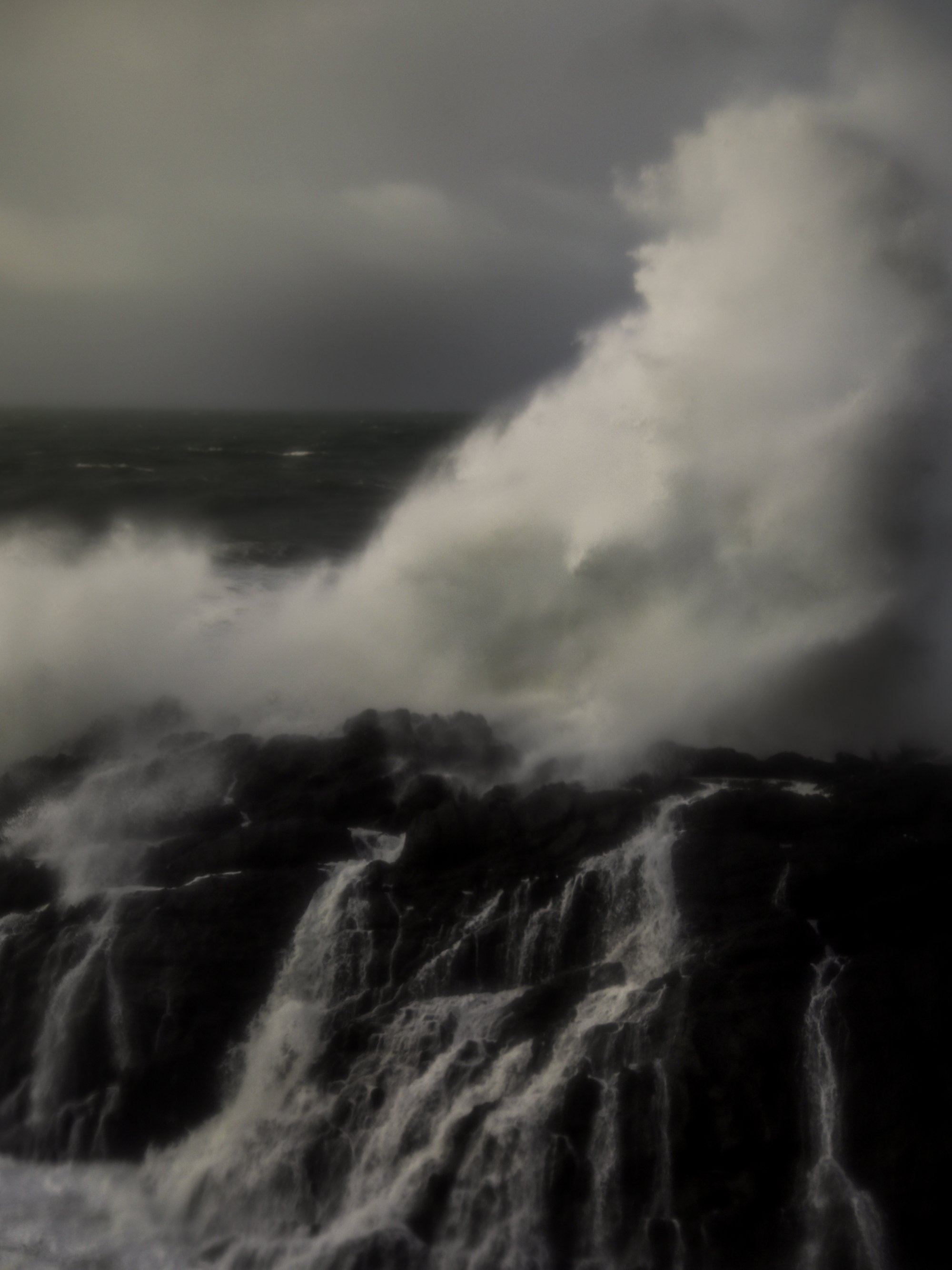 Ocean waves crashing onto dark, rocky shoreline during stormy weather with heavy clouds.