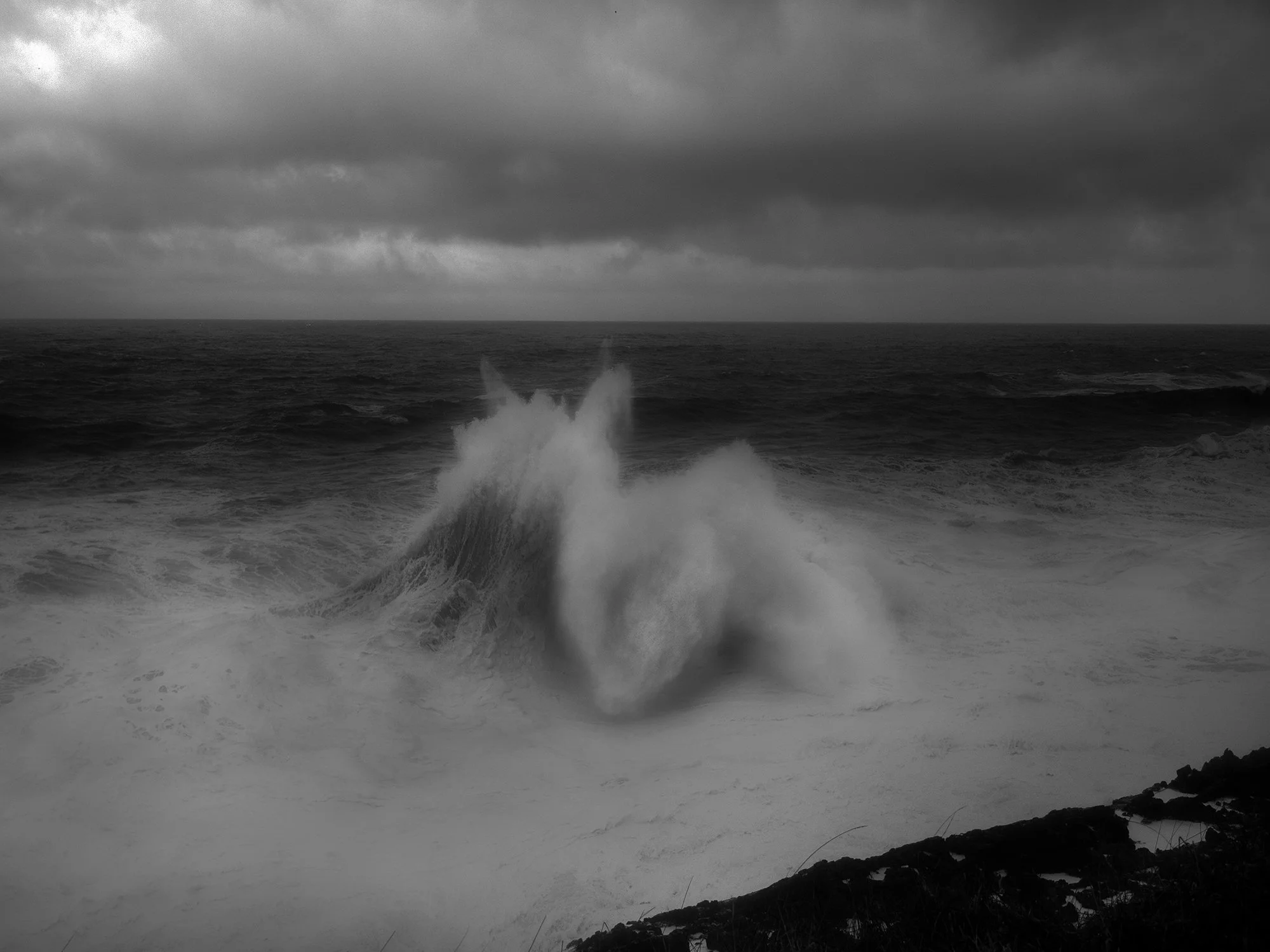 Black and white photograph of the ocean showing large waves crashing against rocks along the shoreline beneath a cloudy sky.