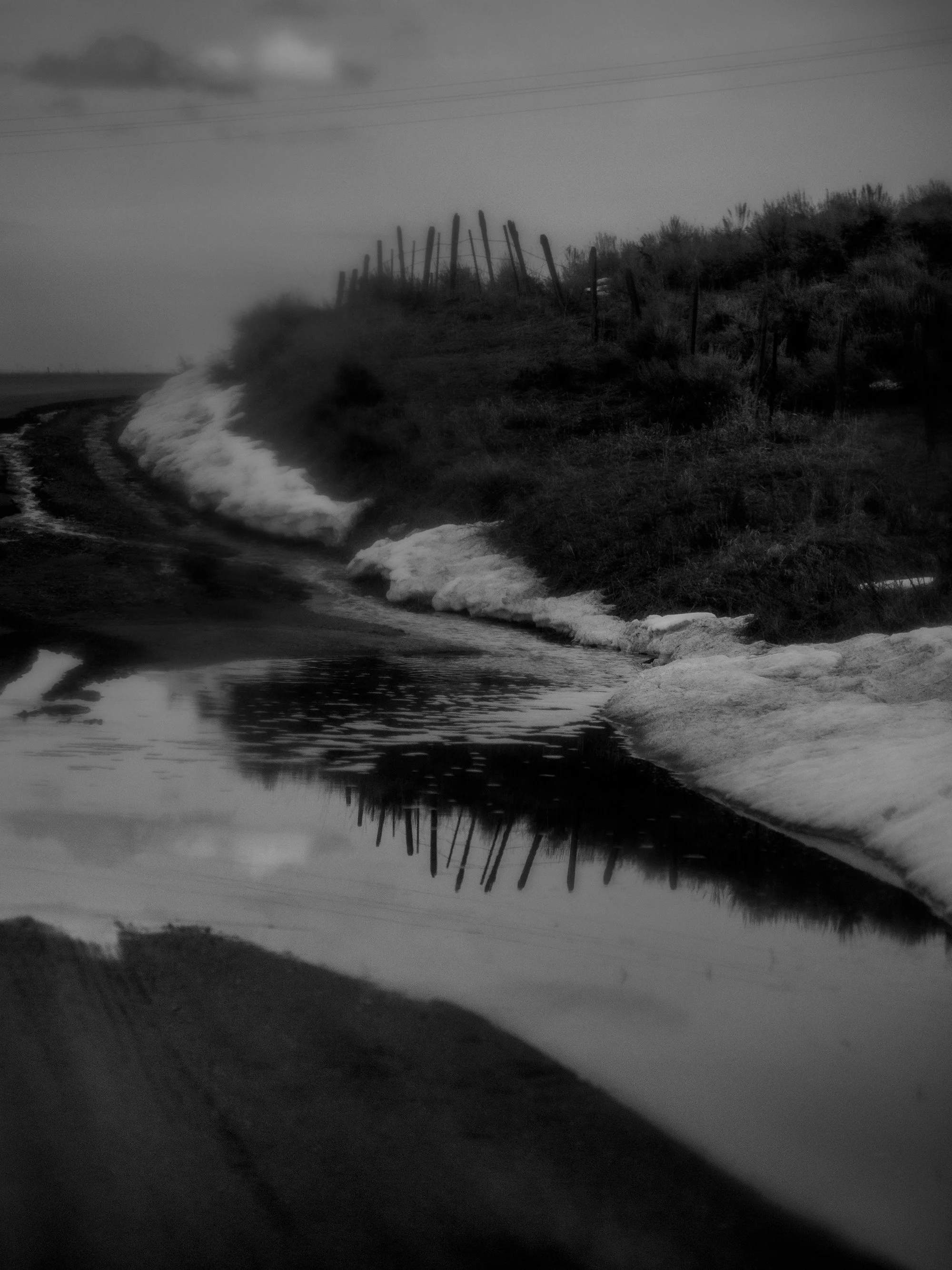 A black and white image of a dirt road with a puddle in the foreground. There is a hill with wooden fence posts and tall grasses on the right side of the image, and utility wires crisscrossing the sky overhead. The scene appears to be rural and quiet