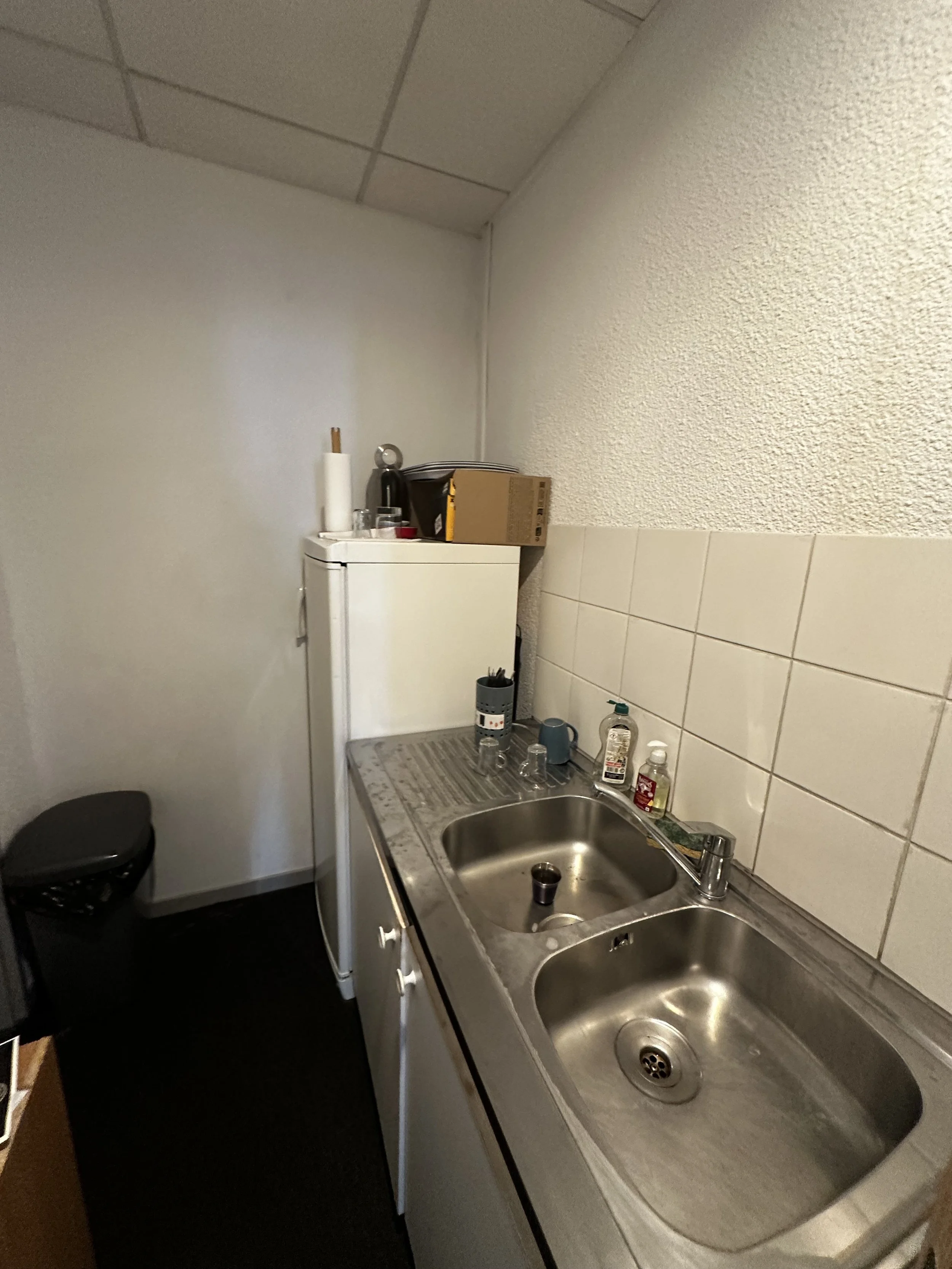 Une petite cuisine avec un évier en acier inoxydable, des couverts, des bouteilles de savon et quelques objets sur le comptoir, mur blanc avec carrelage et un mini réfrigérateur blanc.
