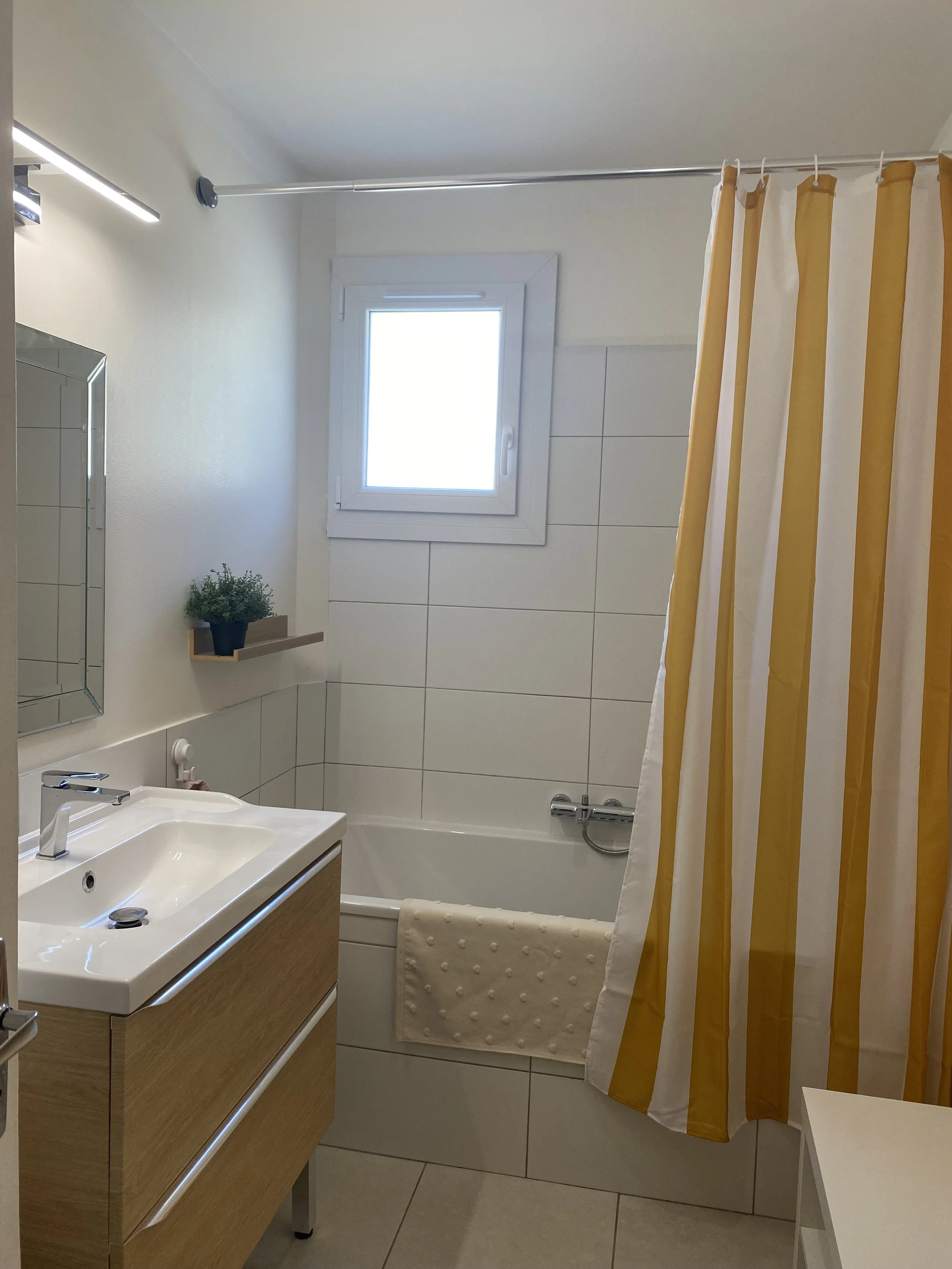 Petite salle de bain avec une baignoire, un lavabo avec un meuble en bois clair, une fenêtre, un miroir, un porte serviette avec une serviette beige, un rideau de douche rayé jaune et blanc, une petite étagère avec une plante verte, et un tapis de bain beige.