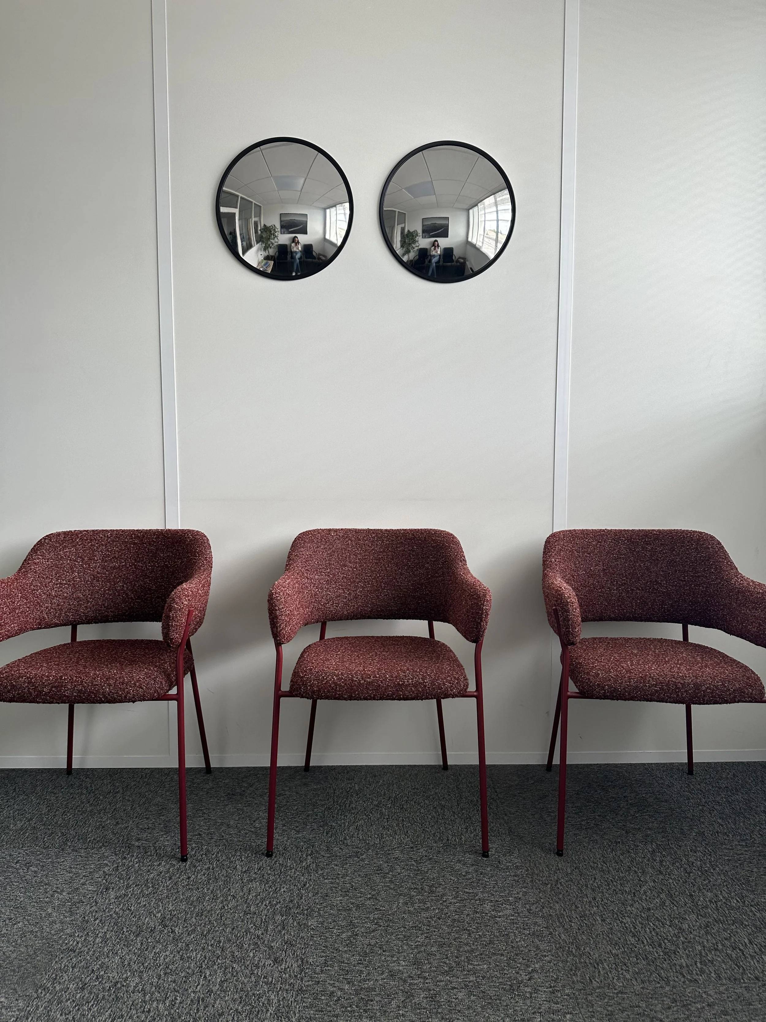 Trois fauteuils rouges alignés contre un mur blanc dans une salle d'attente, avec deux miroirs ronds au-dessus qui reflètent des fenêtres et un cadre noir.