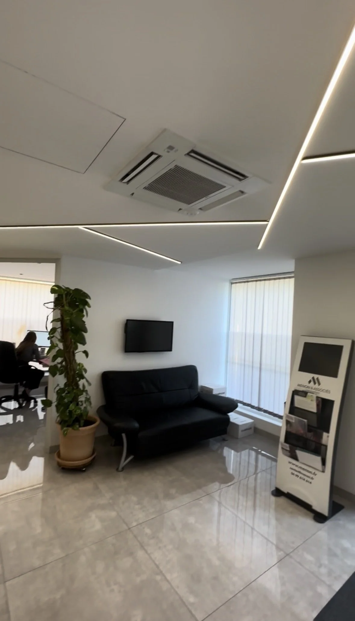 Salle d'attente dans un cabinet médical avec un canapé noir, une plante, une télévision murale et un support de brochures. Le plafond a des lumières encastrées.
