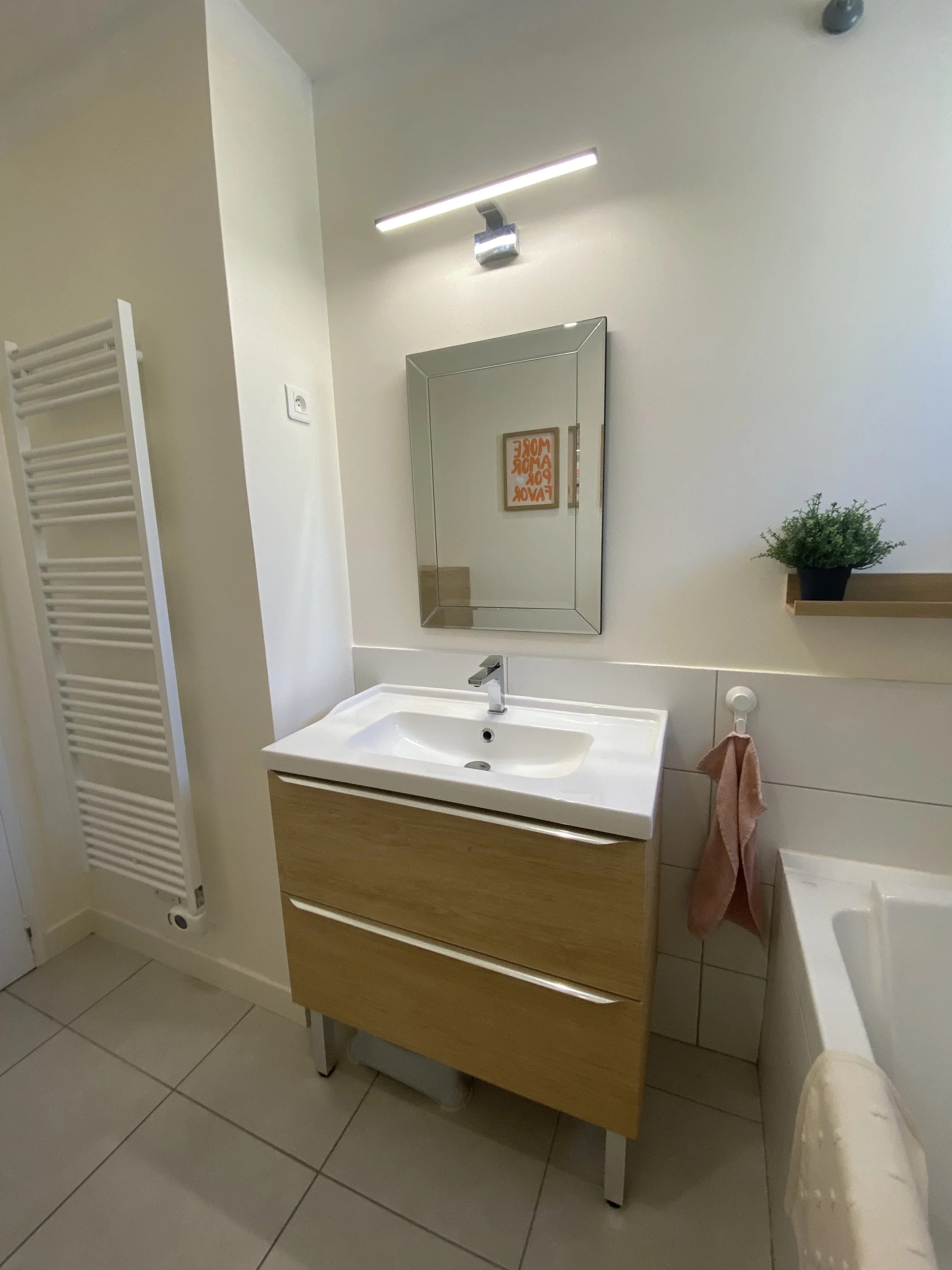Un miroir carré au-dessus d'un lavabo blanc avec meuble en bois clair, à côté d'une étagère en bois avec une plante verte en pot. Il y a une serviette rose suspendue et un sèche-serviette blanc à l'extrémité gauche.