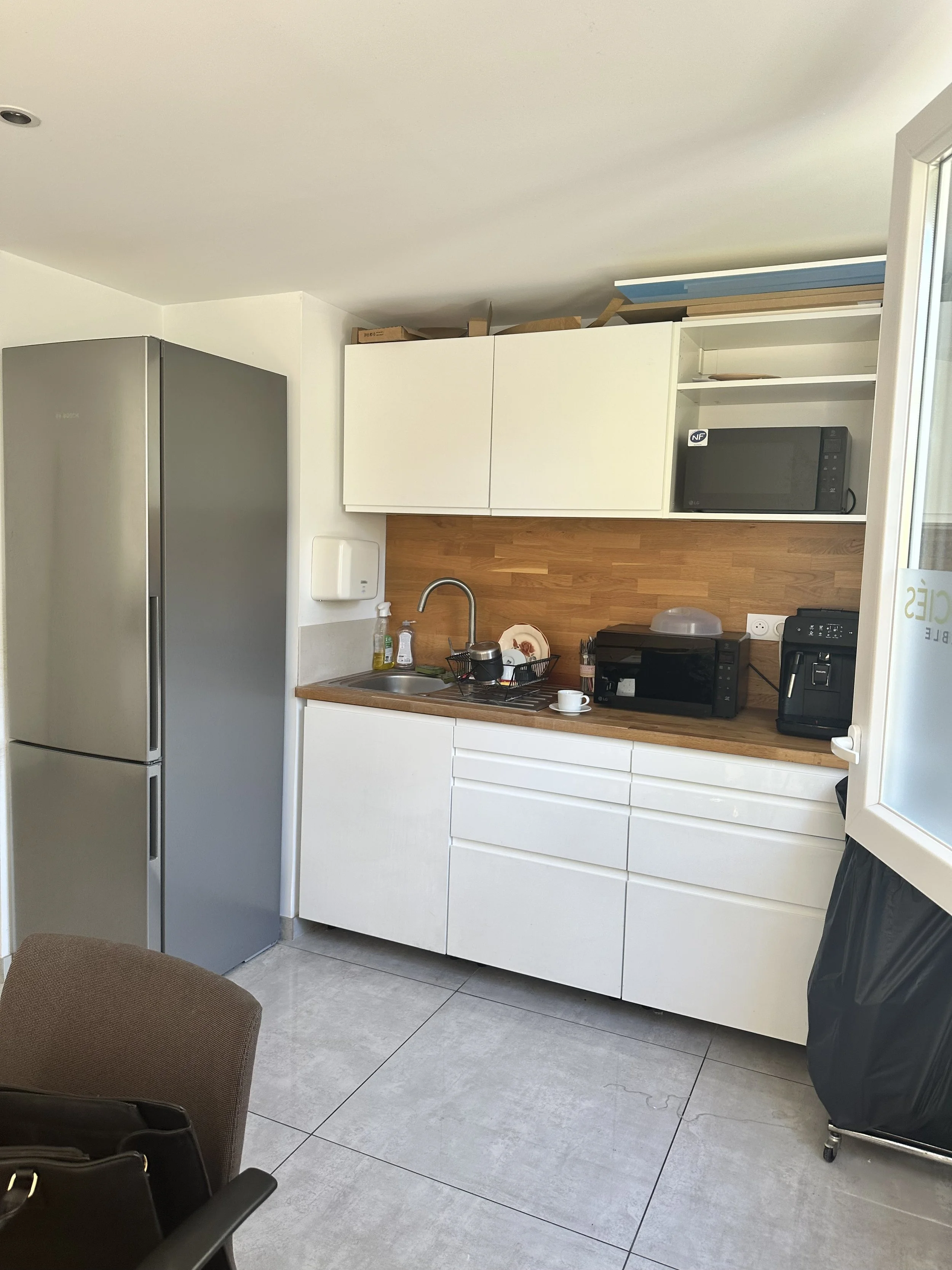 Une cuisine moderne avec un réfrigérateur gris, des armoires blanches, un plan de travail en bois, un évier, une micro-ondes, un grille-pain, une machine à café, un savon liquide, une poubelle en plastique, et un fauteuil marron.