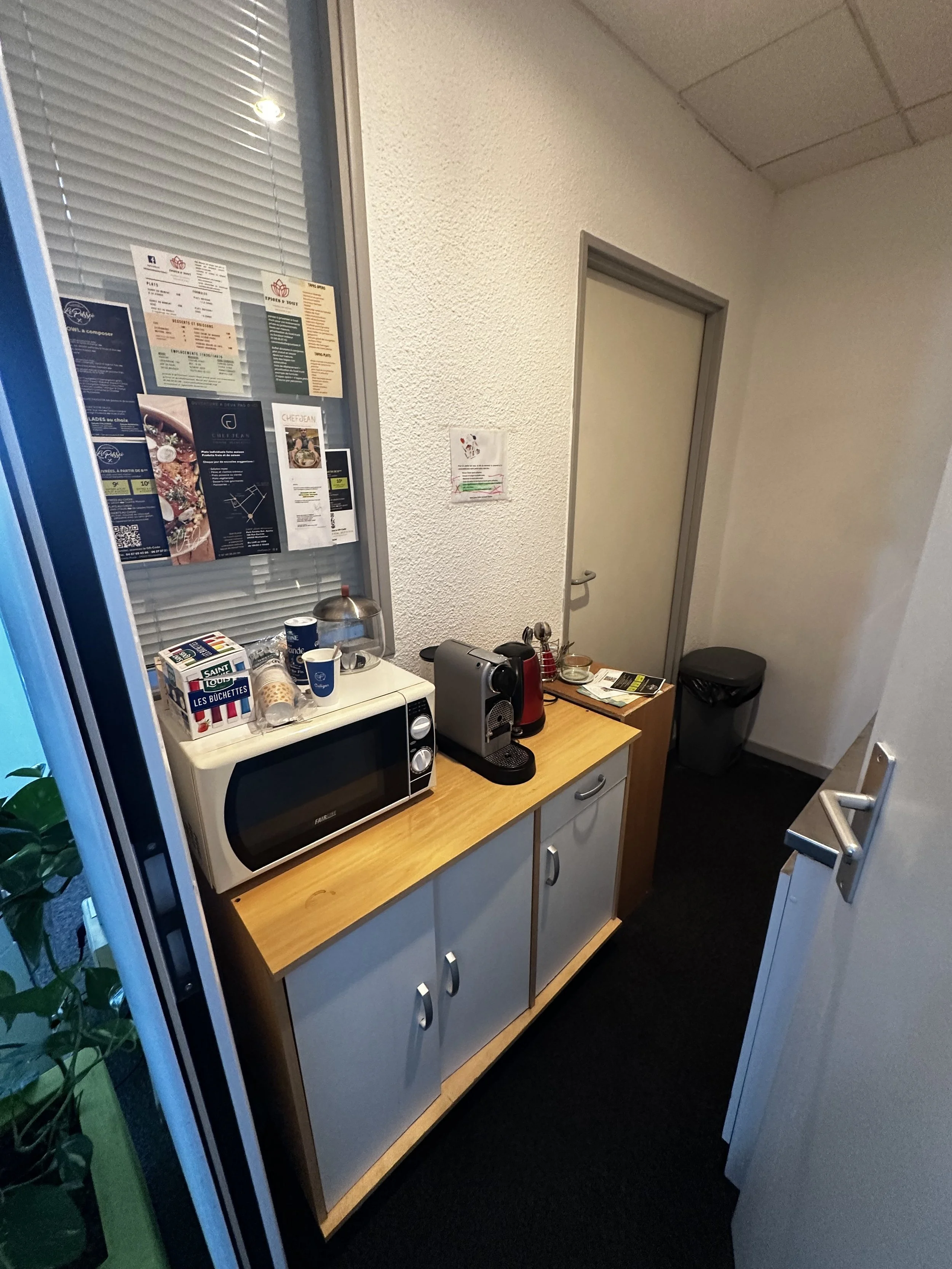 Coin cuisine avec micro-ondes, machine à café, boîtes, papiers, et un tableau de messages ou d'informations sur le mur.