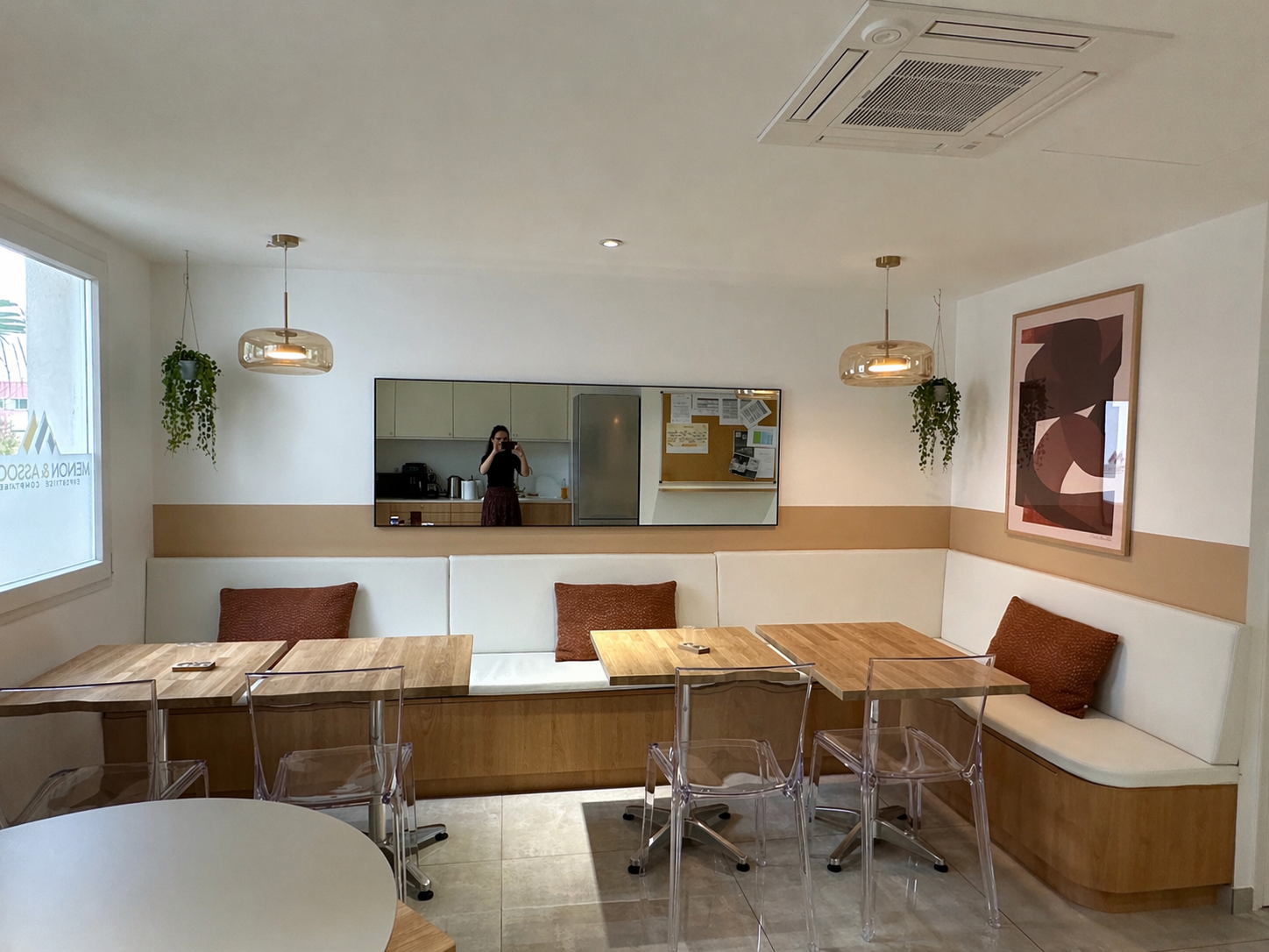 Intérieur d'un café ou restaurant avec des tables en bois, des chaises en plastique transparent, un coin avec des coussins marron, une grande fenêtre, un miroir rectangulaire, une peinture abstraite sur le mur, et des plantes suspendues