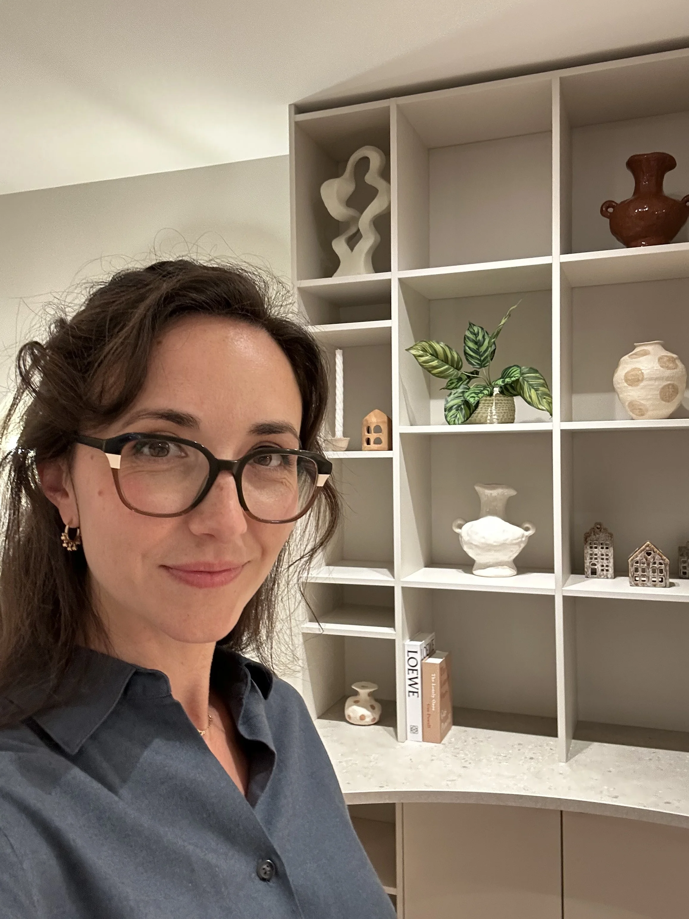 Portrait d'une femme avec lunettes, souriante, portant une chemise noire, posant devant une étagère blanche remplie de décorations, de livres et de plantes.