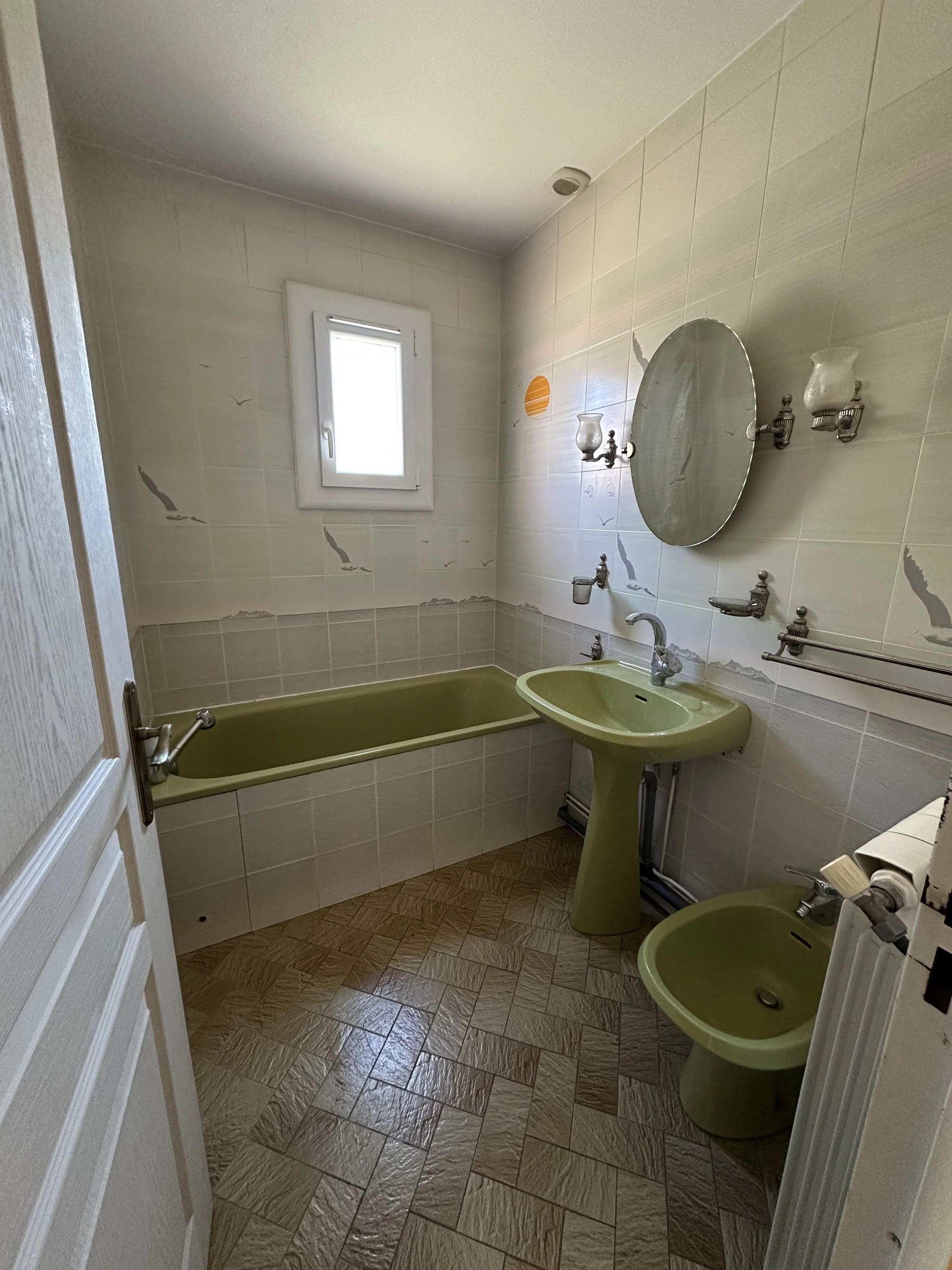 Vue d'une petite salle de bain avec une baignoire vert olive, un lavabo à l'évier de la même couleur, un miroir rond, des appliques murales en verre, un petit cabinet en dessous du lavabo, une fenêtre en haut du mur, des carreaux de faïence blanche avec des motifs d'oiseaux en vol. Le sol est en carrelage marron avec un motif en carreaux.