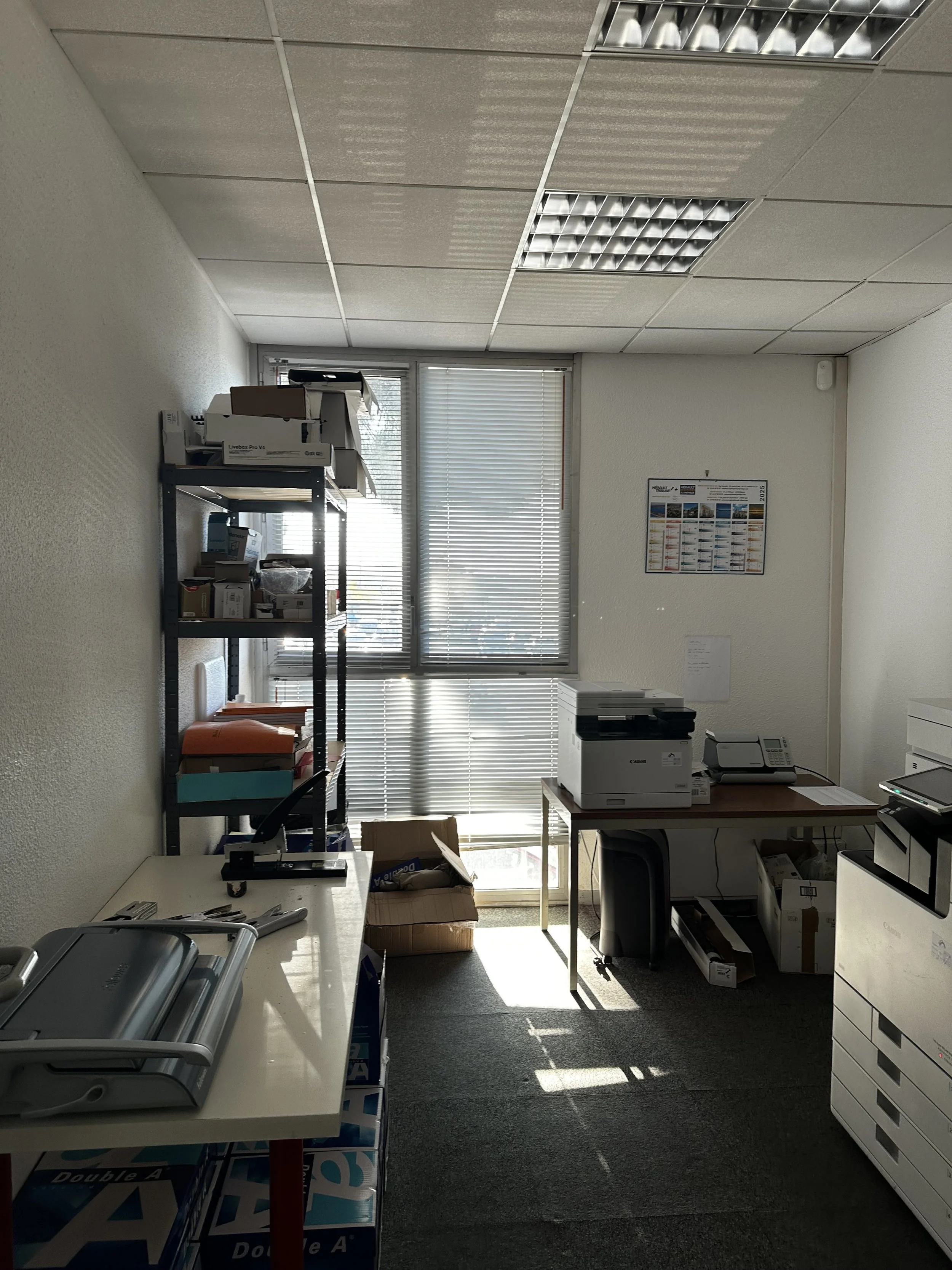 Un bureau avec des étagères contenant des boîtes, un photocopieur, une table avec une imprimante et une boîte en carton. La pièce est éclairée par la lumière du soleil à travers des stores vénitiens.
