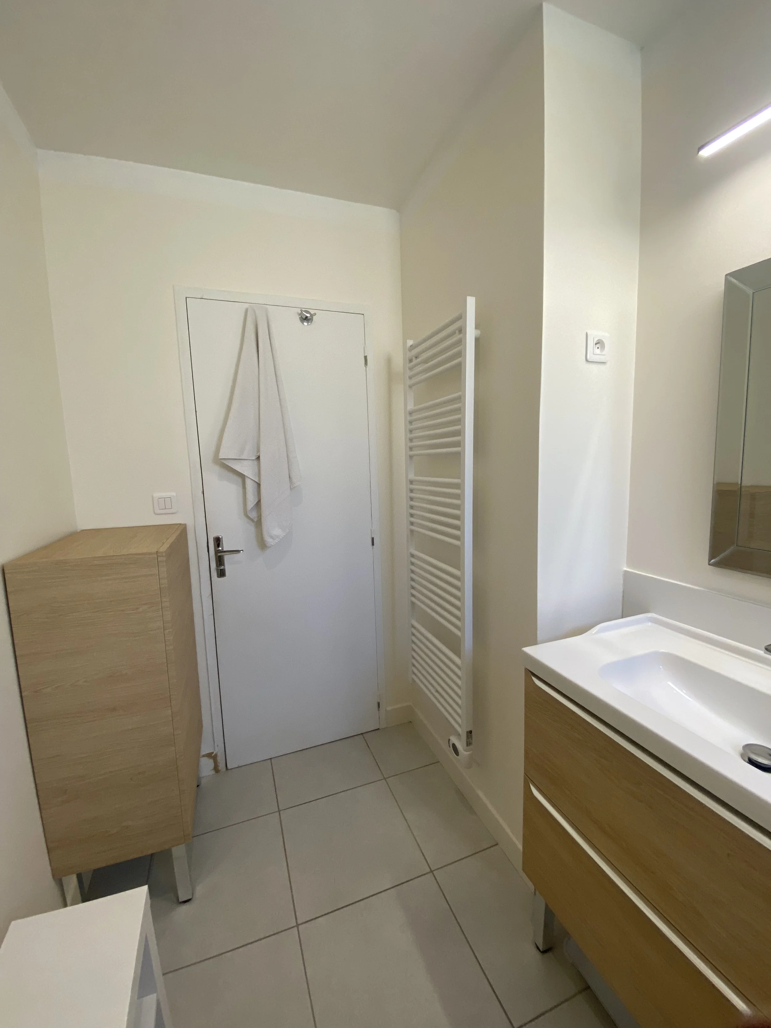 Intérieur d'une salle de bain avec un lavabo en bois clair, un miroir, un radiateur blanc, une porte blanche avec une serviette blanche suspendue, et un meuble en bois clair placé sous le lavabo.