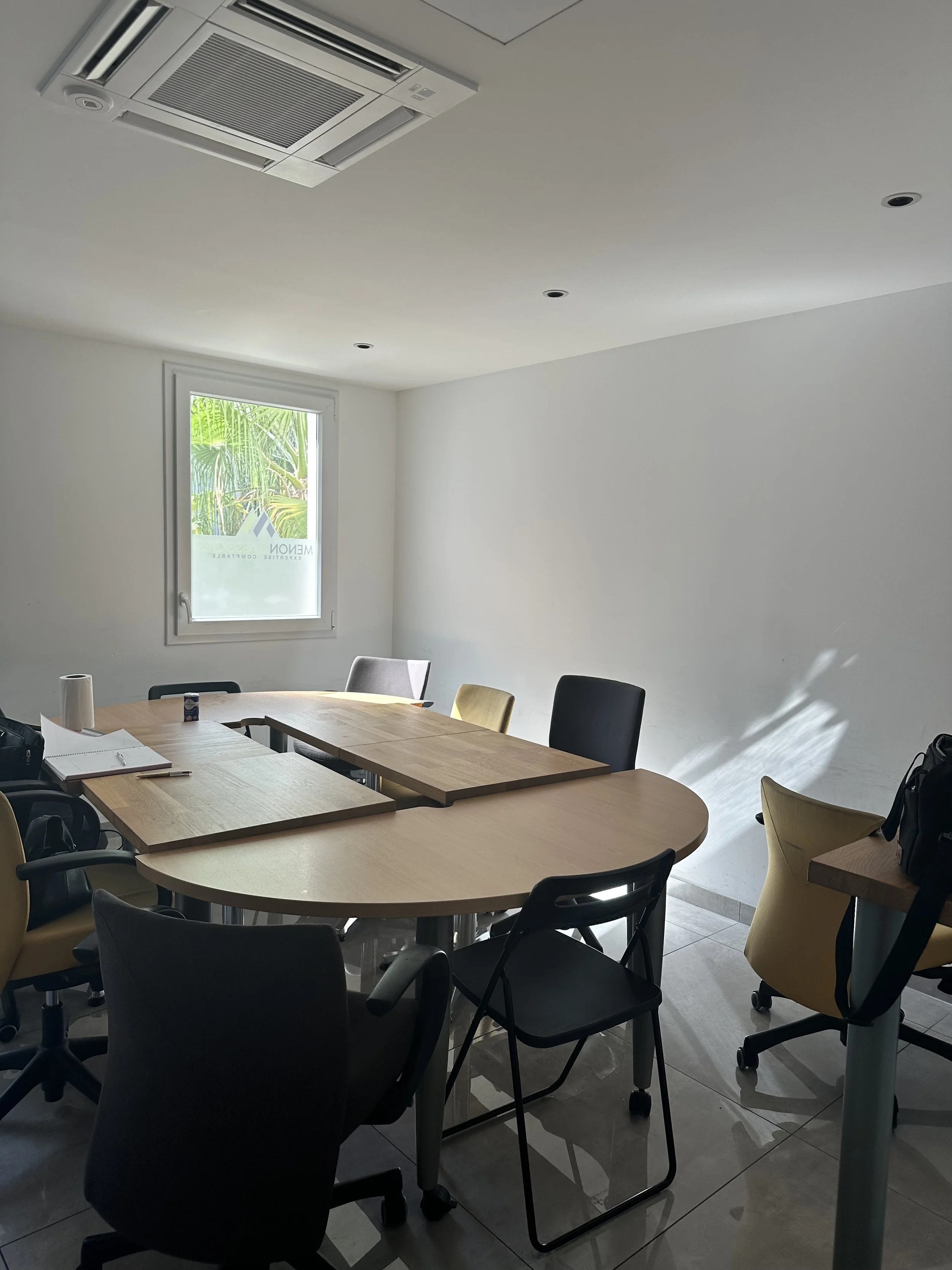 Salle de réunion avec une table en bois et plusieurs chaises autour, une fenêtre laissant entrer la lumière naturelle, un rouleau de papier toilette, un carnet, et un sac noir présents sur la table.