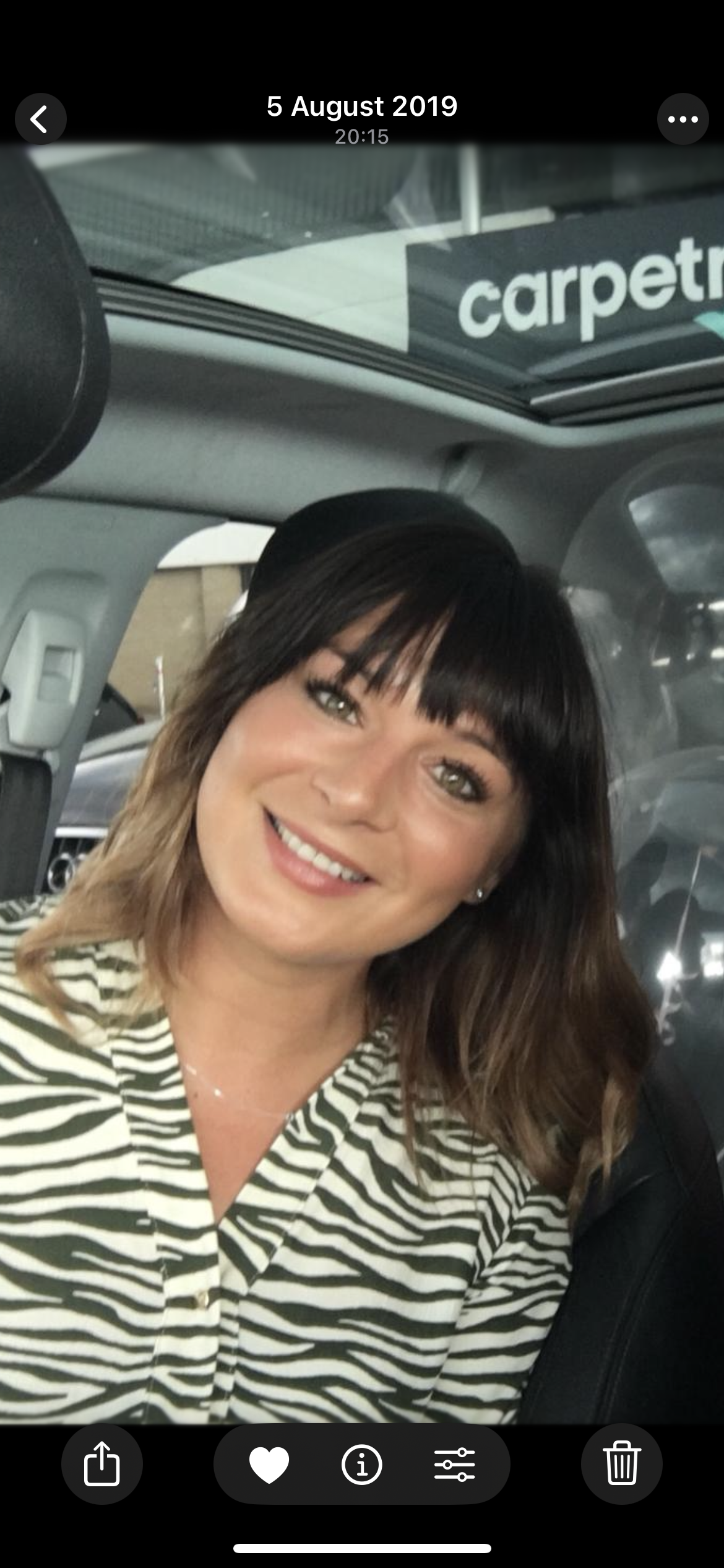 A smiling woman with shoulder-length brown hair and bangs, wearing a zebra-patterned shirt, sitting inside a vehicle.