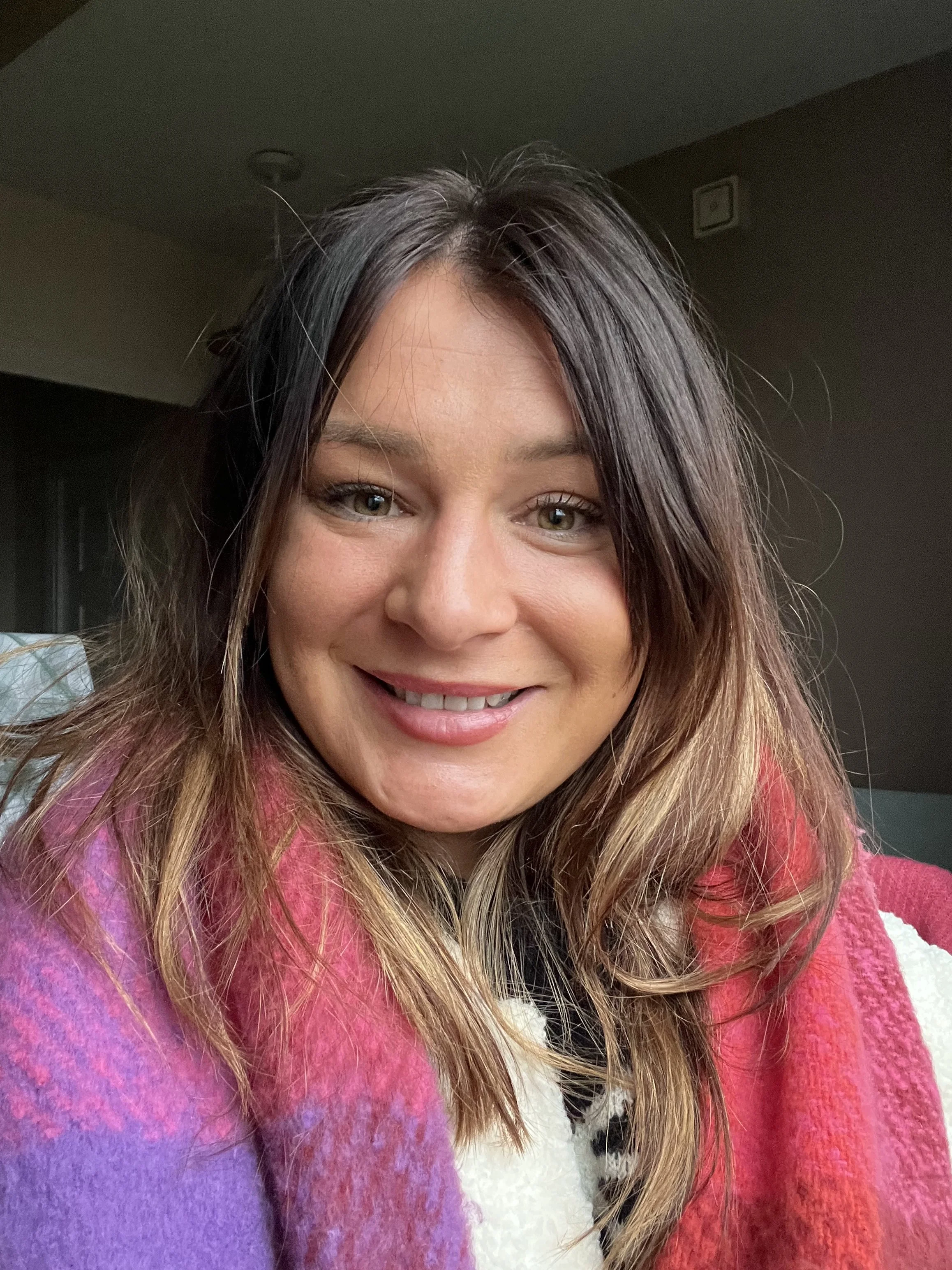 A woman with long, wavy hair smiling, wearing a colorful pink and purple scarf, and a white and black patterned top.