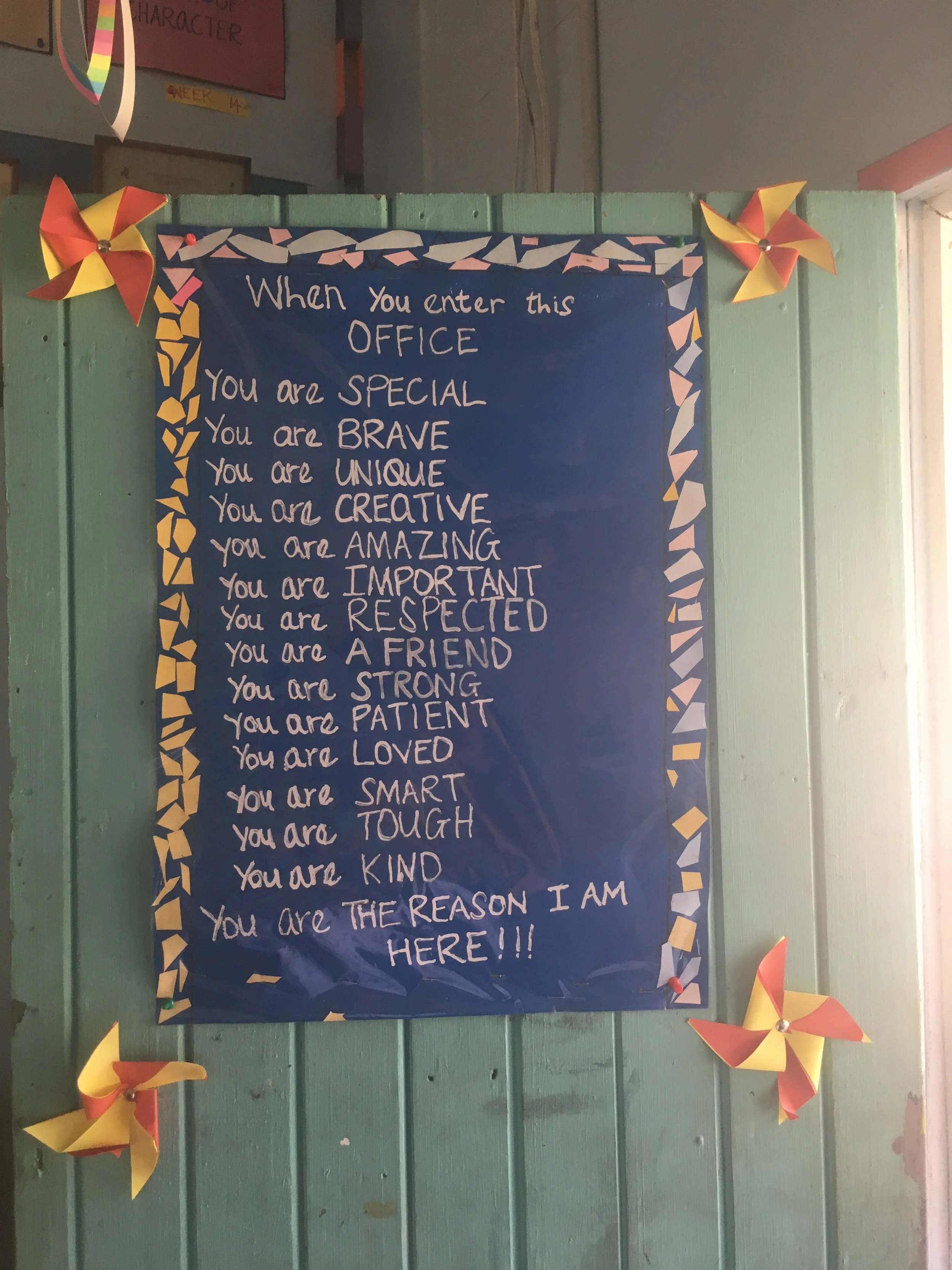 A decorated bulletin board with a motivational message in white letters on a dark blue background. The message reads: 'When you enter this office, you are special, brave, unique, creative, amazing, important, respected, a friend, strong, patient, loved, smart, tough, kind. The board is bordered with colorful geometric paper, and there are red and yellow paper pinwheels attached to the corners.