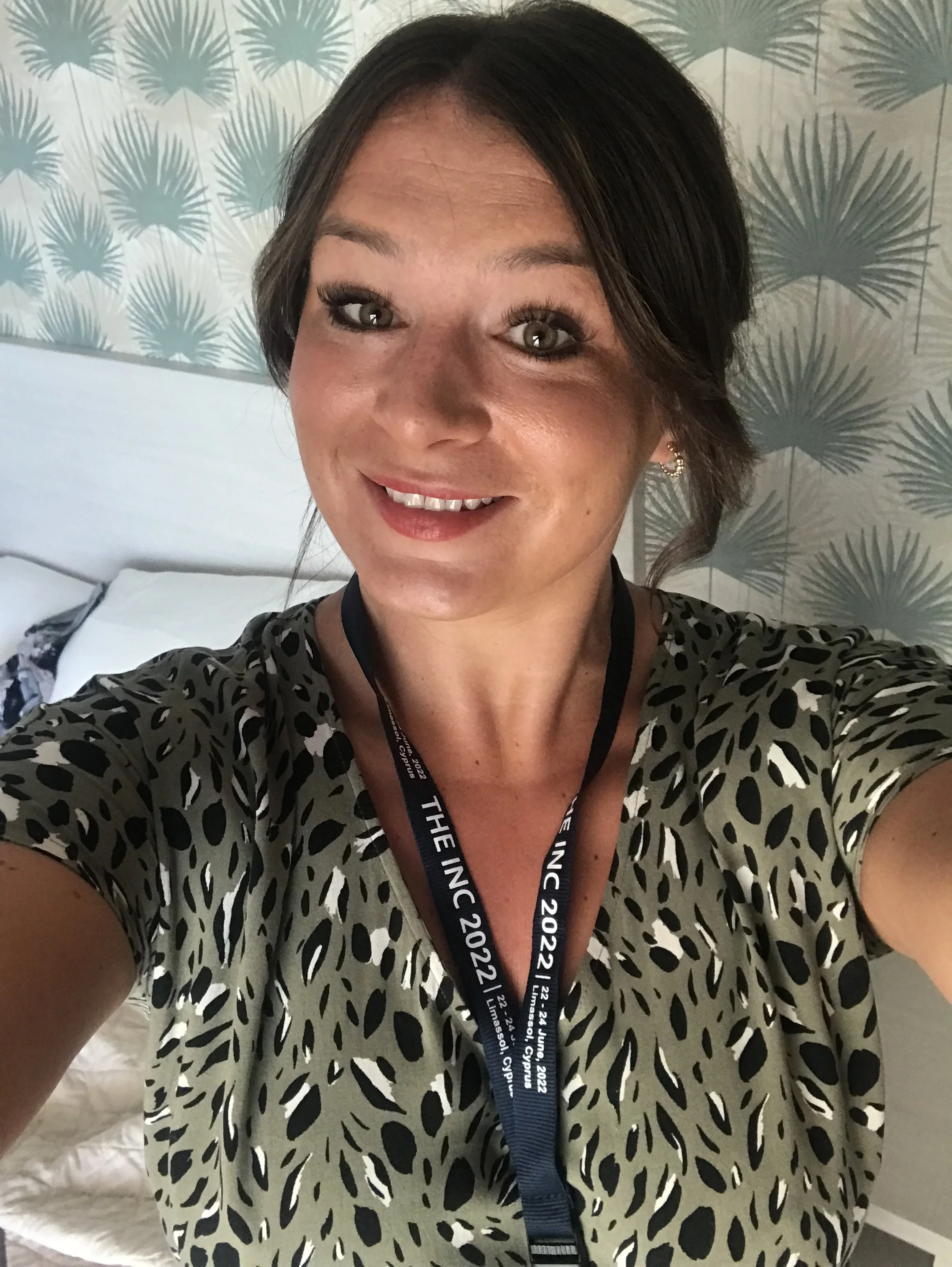 A woman with dark hair styled in a bob taking a selfie. She is wearing a black and white animal print top, earrings, and a conference lanyard around her neck with the text "THE INC 2022". The background features a patterned wallpaper with large palm leaves.
