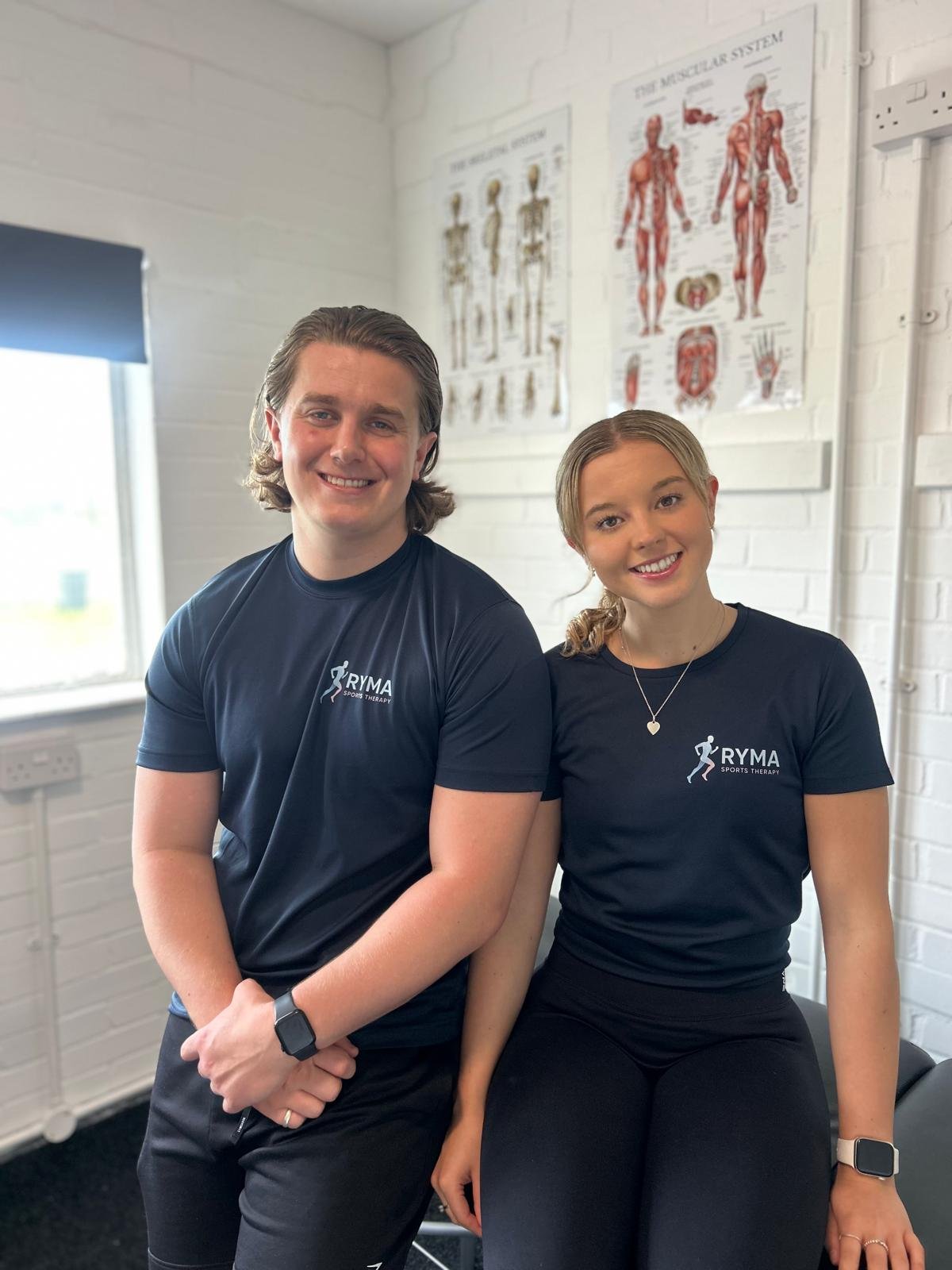 Two smiling young adults, male and female, wearing navy blue sports therapy shirts with 'RYMA SPORTS THERAPY' logo, sitting in a room with white brick walls and anatomical posters, posing confidently.