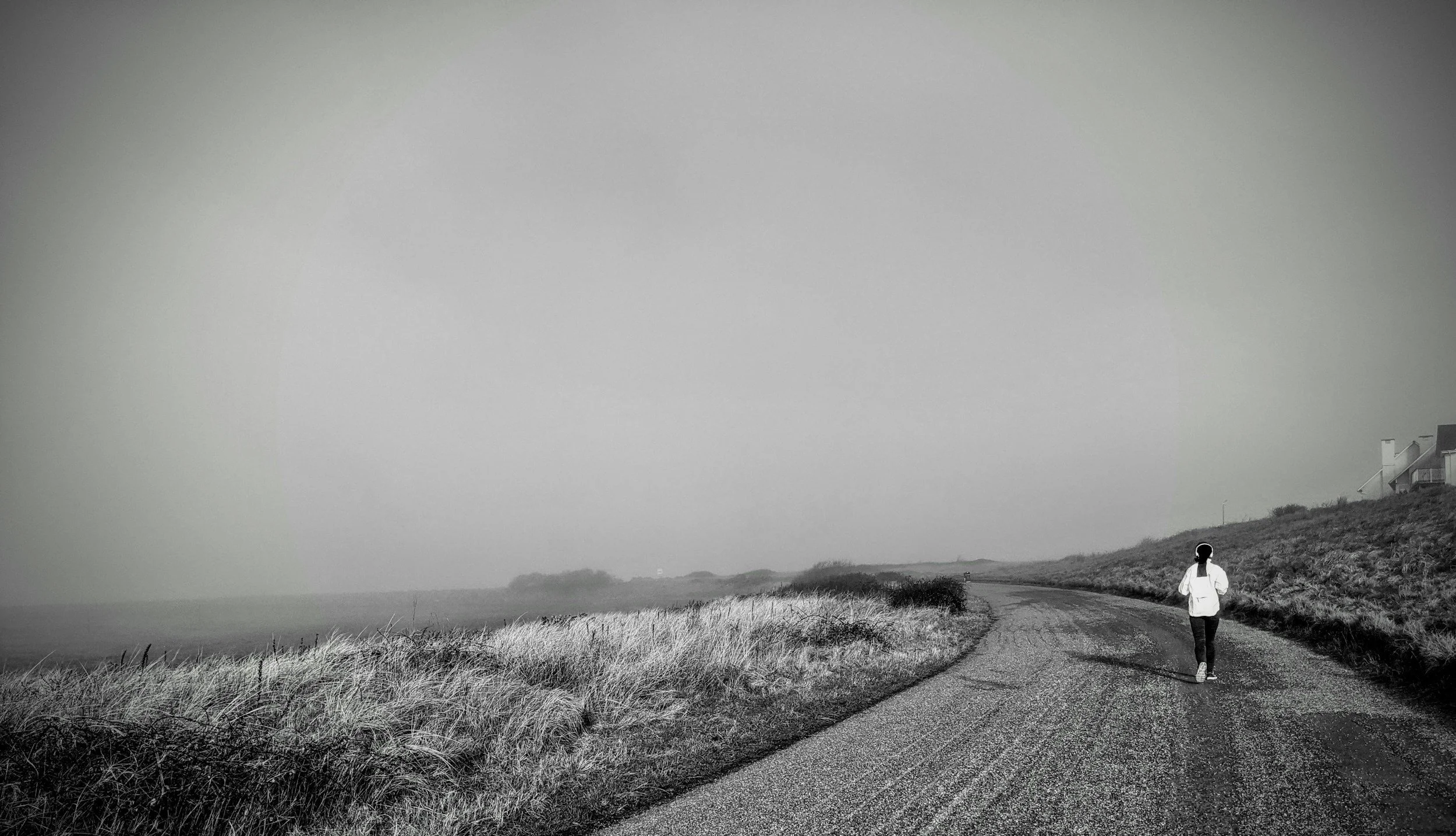 A person walking along a dirt path near the coast with grassy dunes on the left and houses on a hill on the right, under a cloudy sky in black and white.