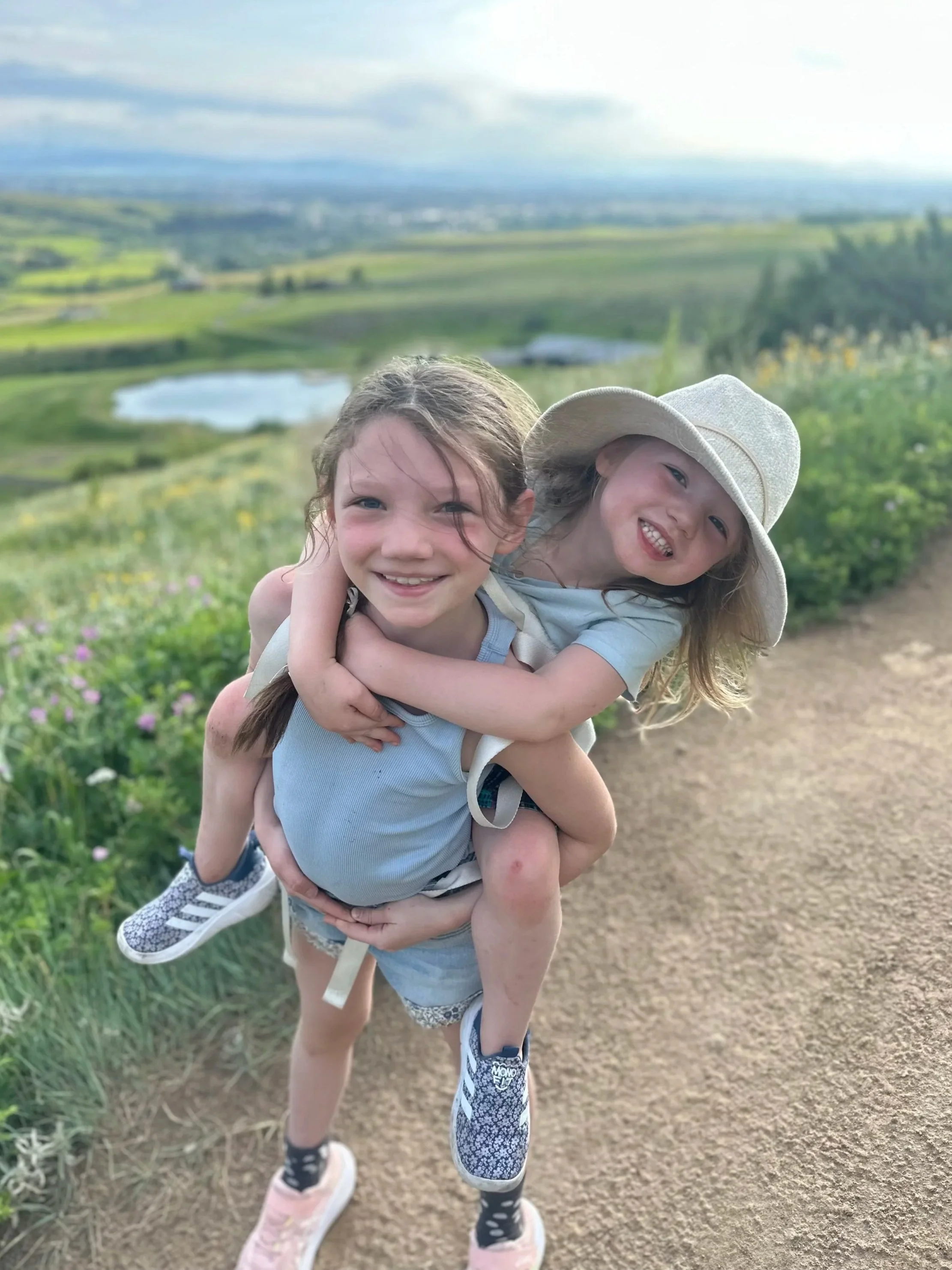 Two young girls smiling and playing outdoors on a trail with a scenic landscape in the background, including green fields and a pond.
