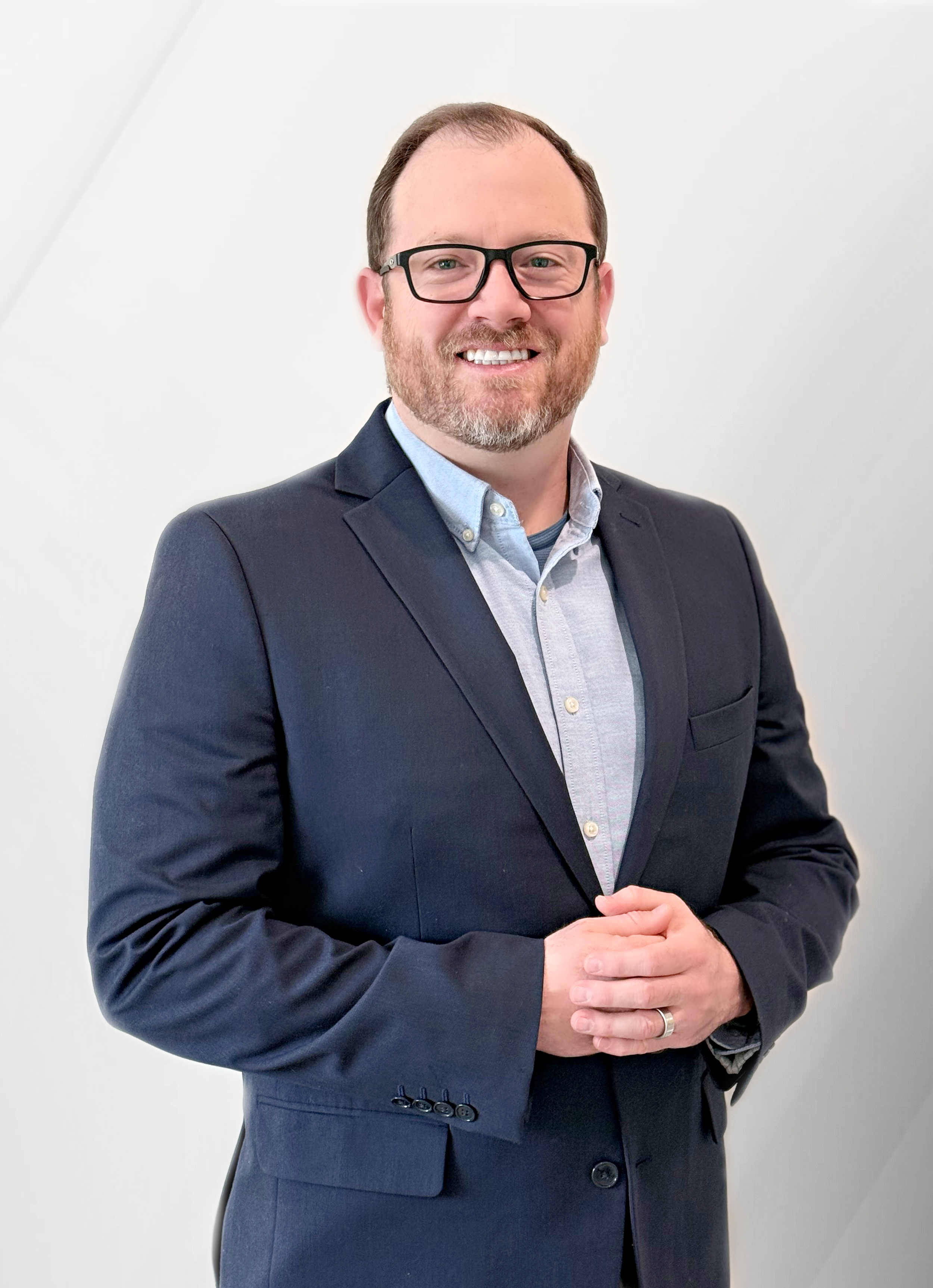 A man with a beard and glasses wearing a dark suit jacket and light blue button-up shirt, smiling and standing against a plain white wall.
