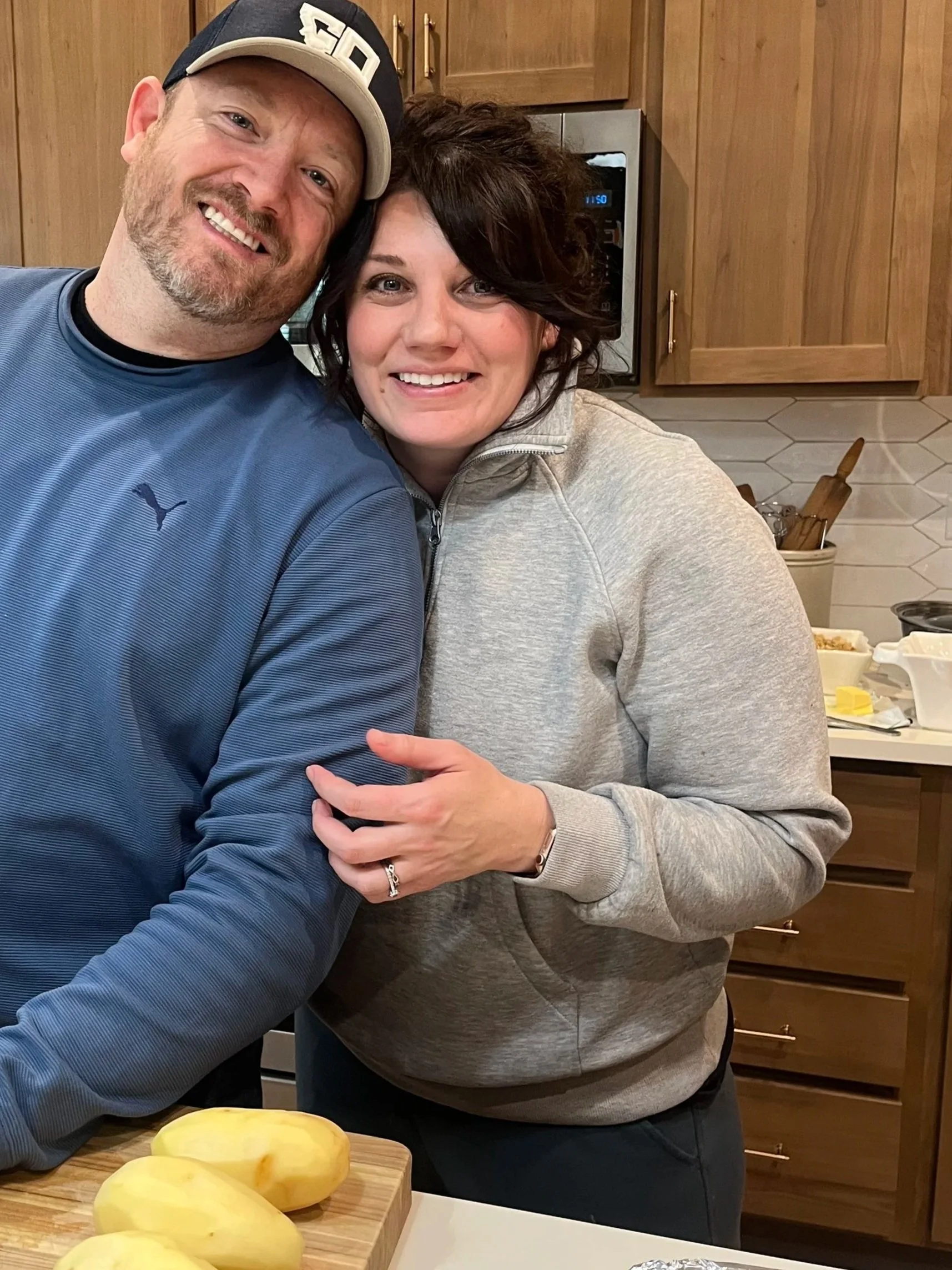 A smiling man wearing a black and white baseball cap and a blue sports shirt, and a smiling woman with short dark hair and a gray zip-up hoodie, standing together in a kitchen. The man hugs the woman, and they are both smiling at the camera. There is a wooden cutting board with yellow vegetables in front of them, and kitchen cabinets and appliances are visible in the background.
