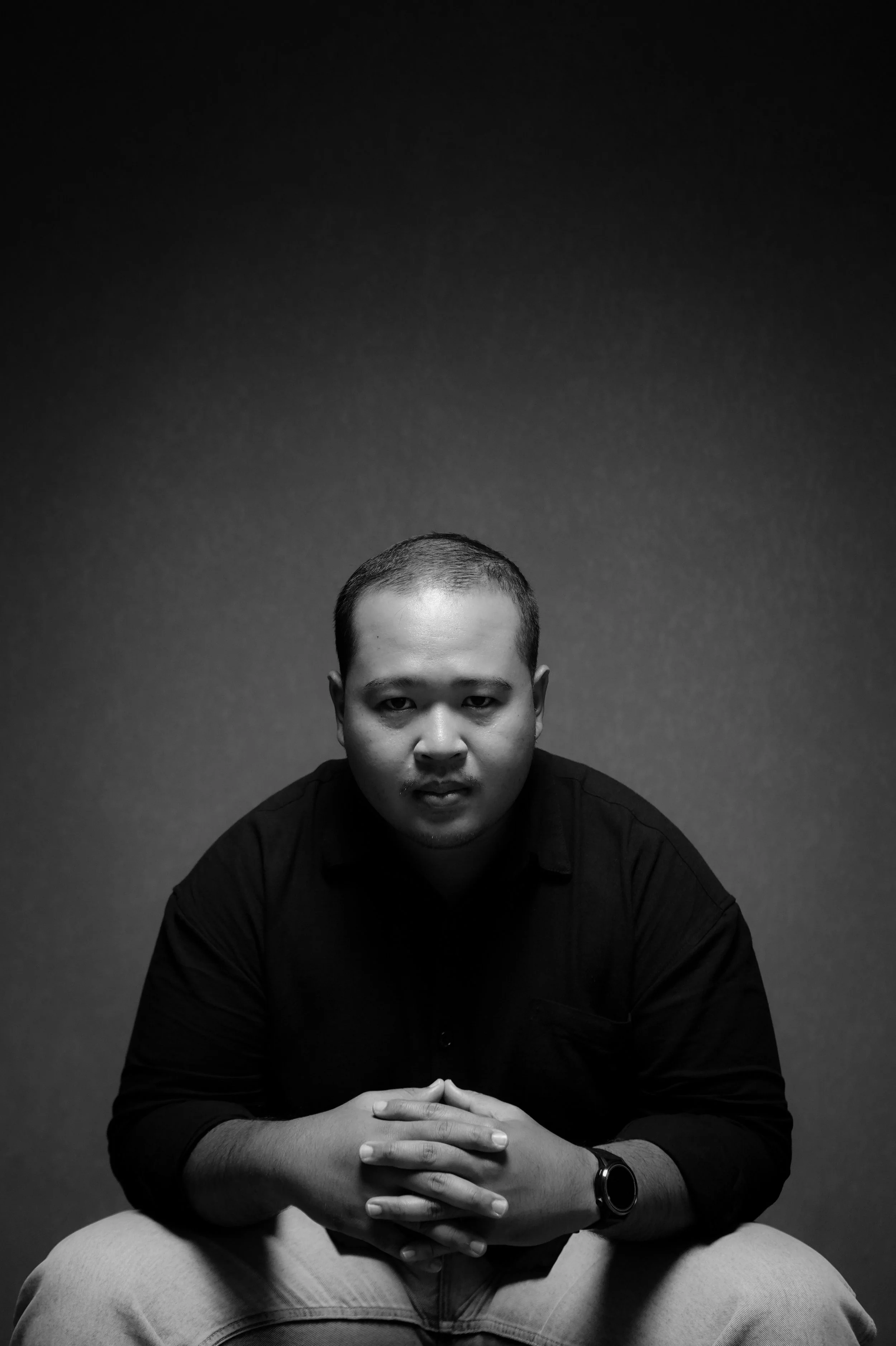 Black and white portrait of a man sitting with his hands clasped, looking directly at the camera, against a dark background.