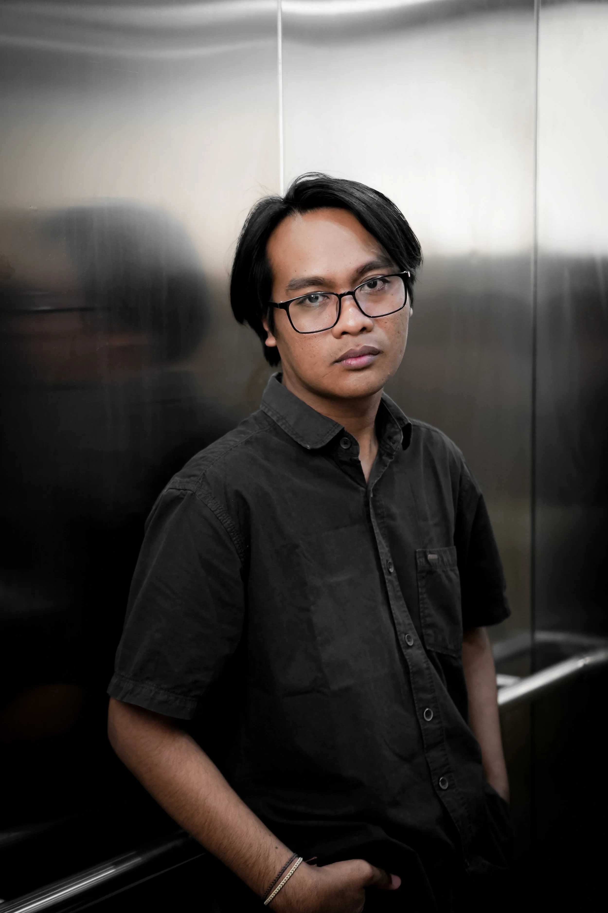 A young man with black hair, glasses, and a black shirt standing in an elevator with metallic walls.