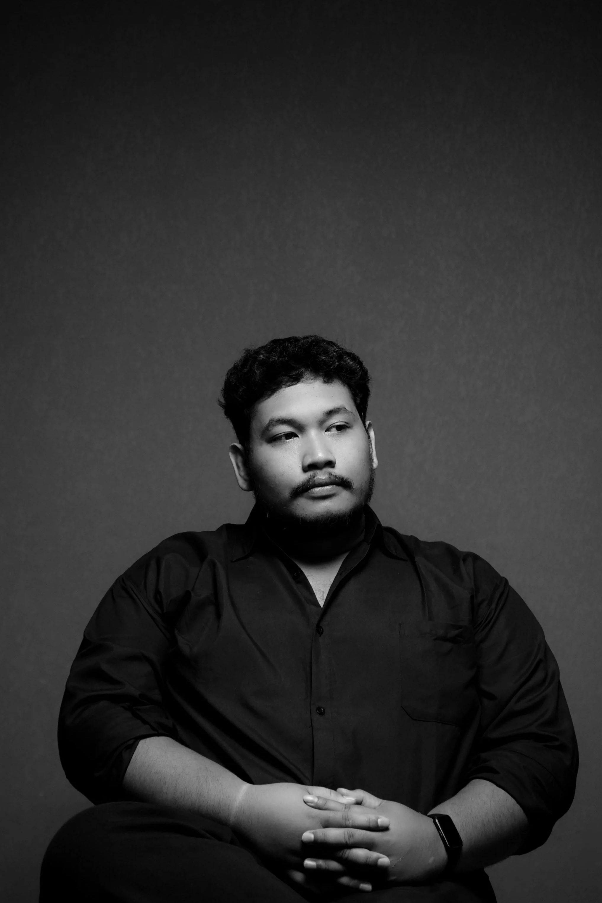 A black-and-white portrait of a man with short dark hair, a mustache, and a beard, wearing a dark shirt, sitting with arms crossed, looking slightly to his right, against a plain dark background.