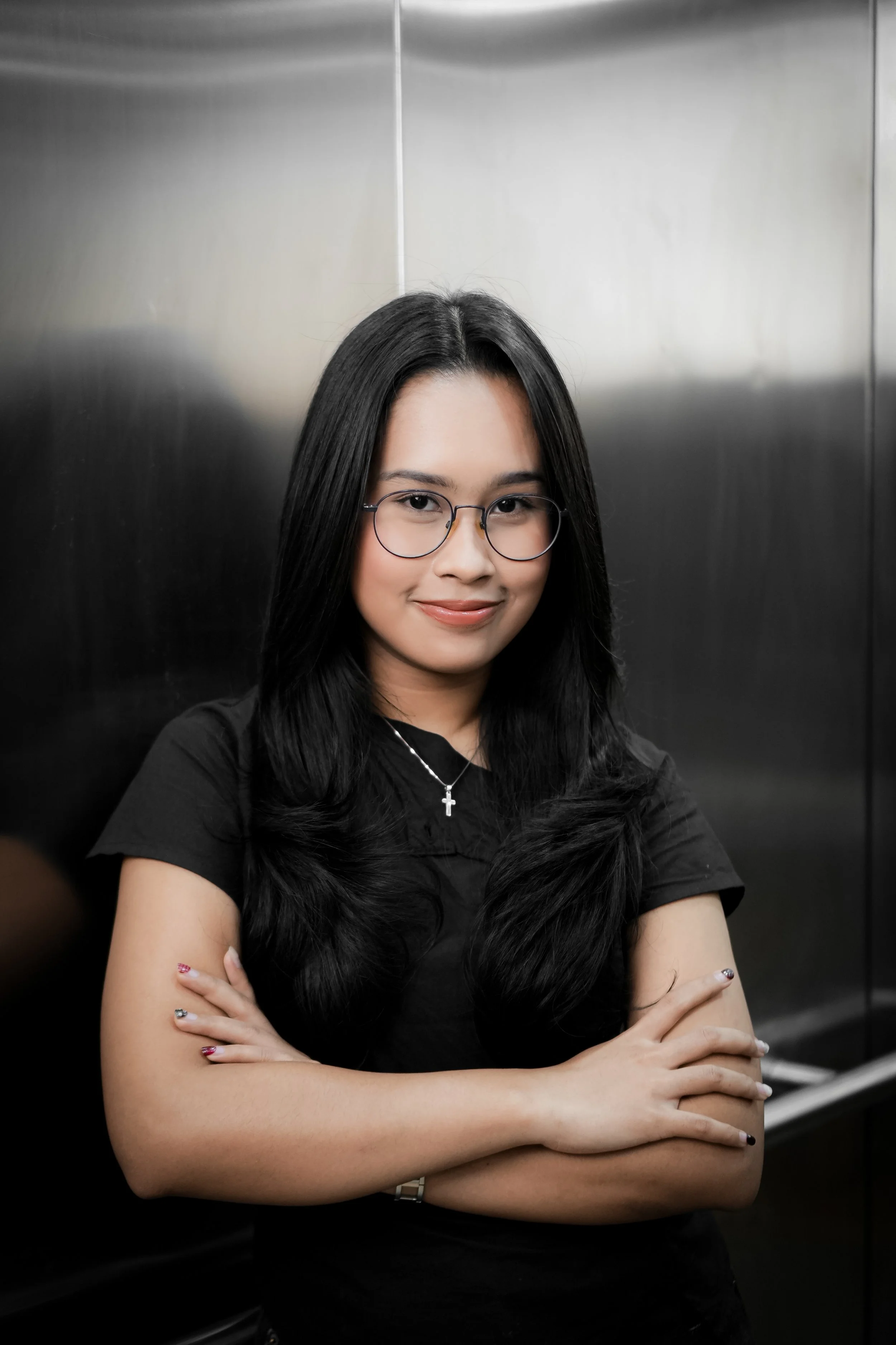 Young woman with long black hair, glasses, and a necklace with a cross, smiling and standing with arms crossed in an elevator with metallic walls.