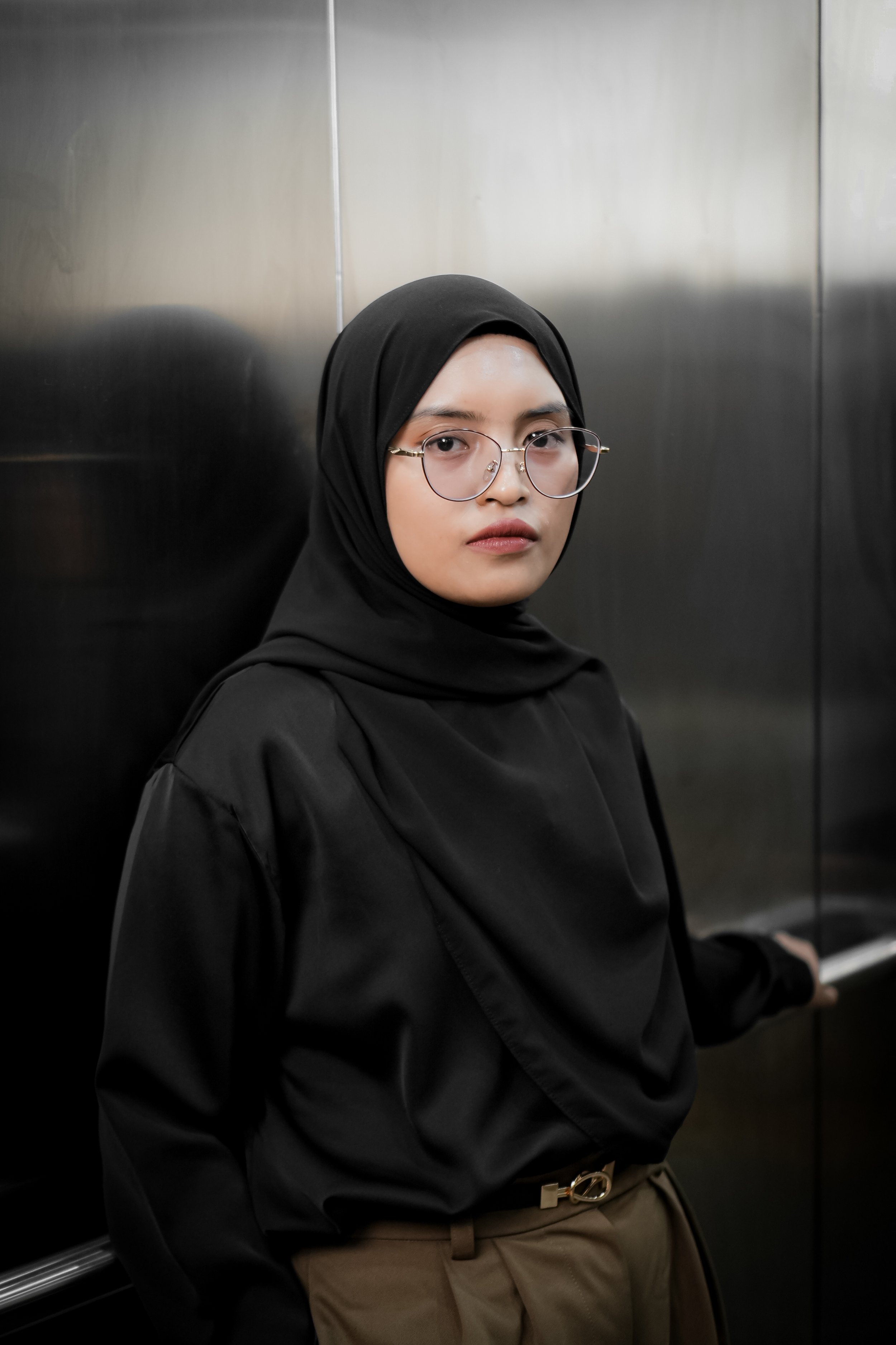 Woman wearing glasses and a black hijab sitting in an elevator with metallic walls.