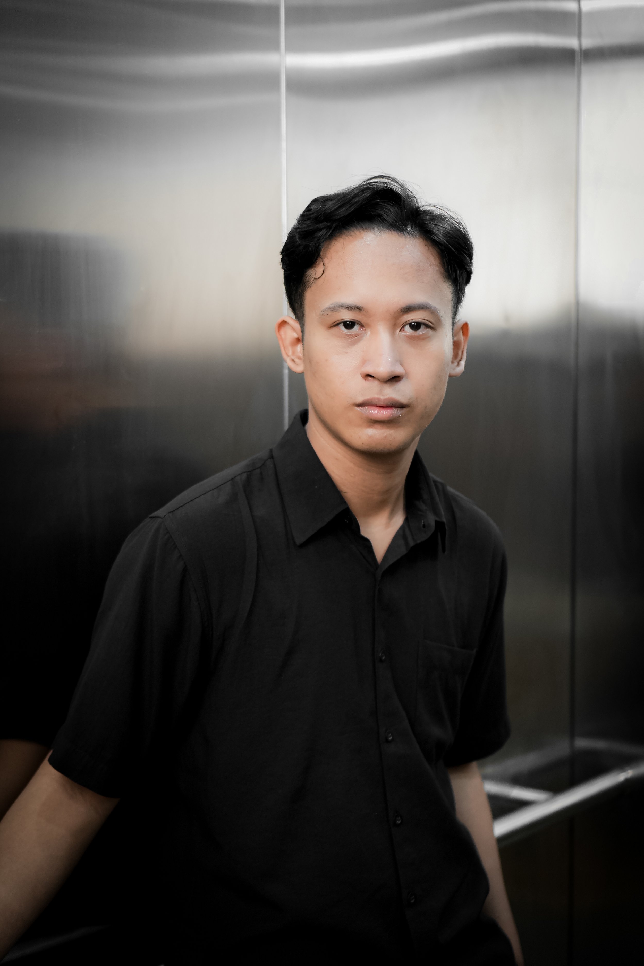 Young man with black hair wearing a black shirt, standing inside an elevator with metallic walls.
