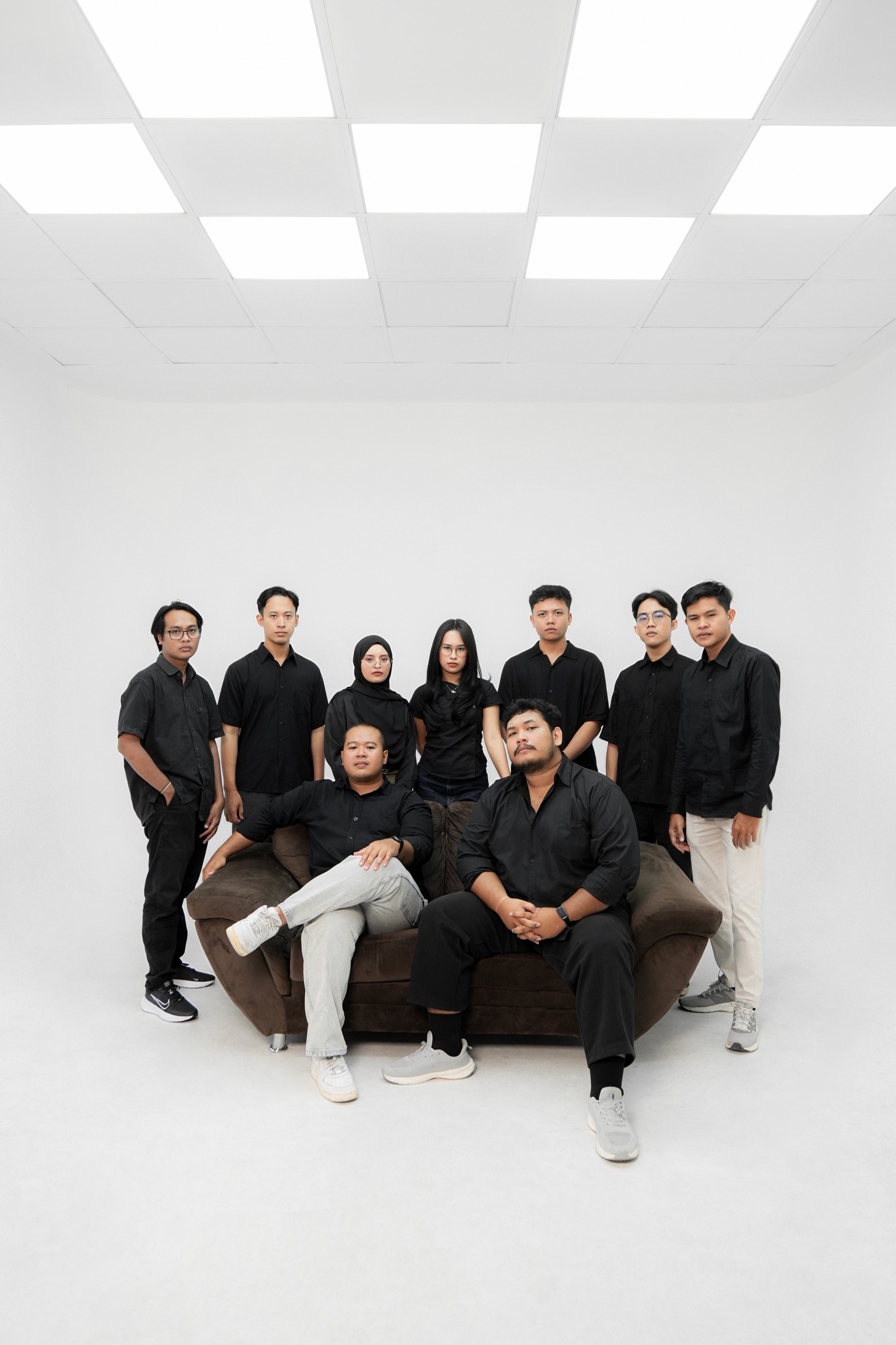 Group of seven people in black shirts standing and sitting around a brown couch in a minimalist room with white walls and ceiling lighting panels.