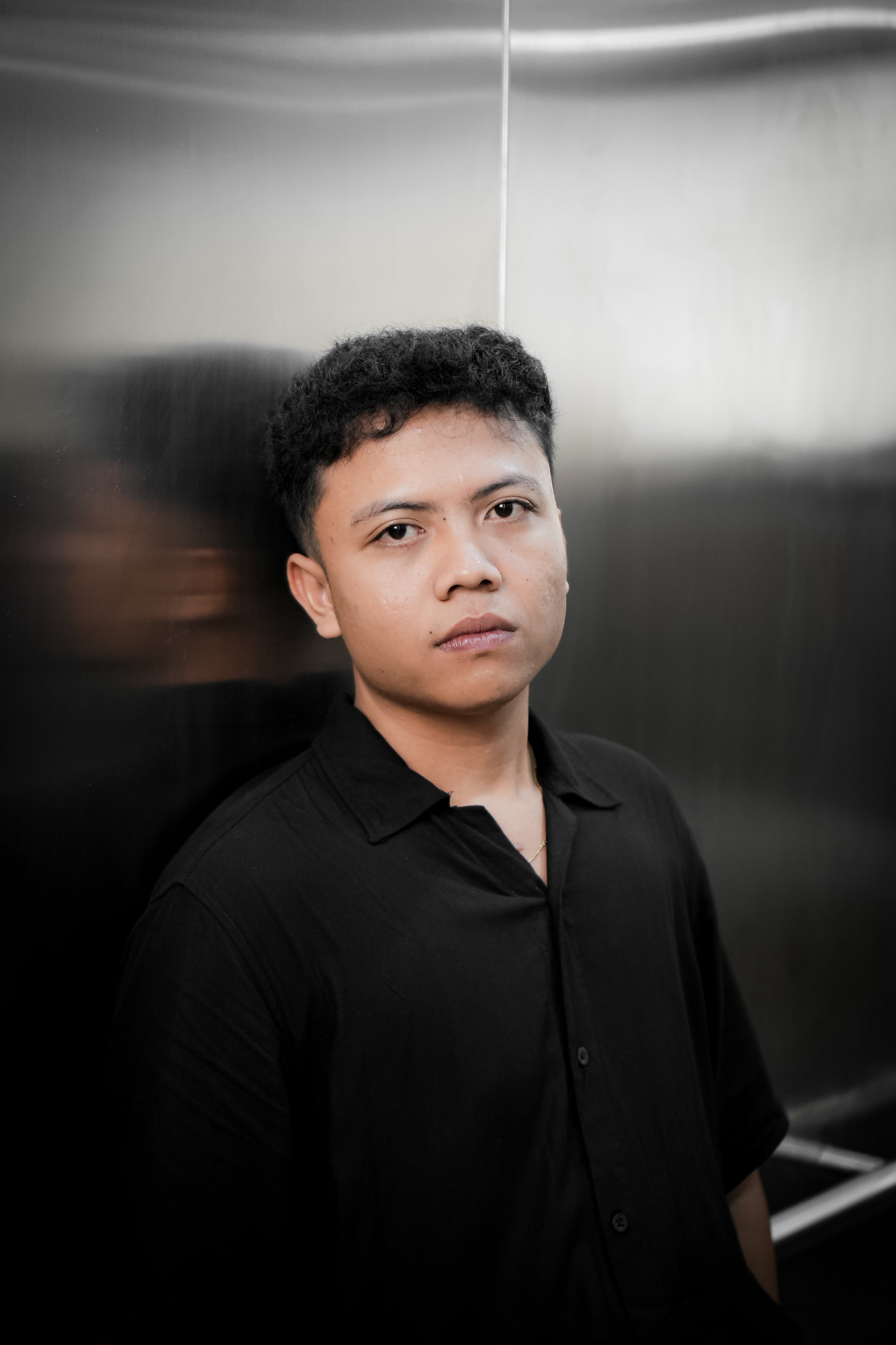 A young man with short, curly black hair and a serious expression, wearing a black collared shirt, standing in front of a metallic elevator wall.