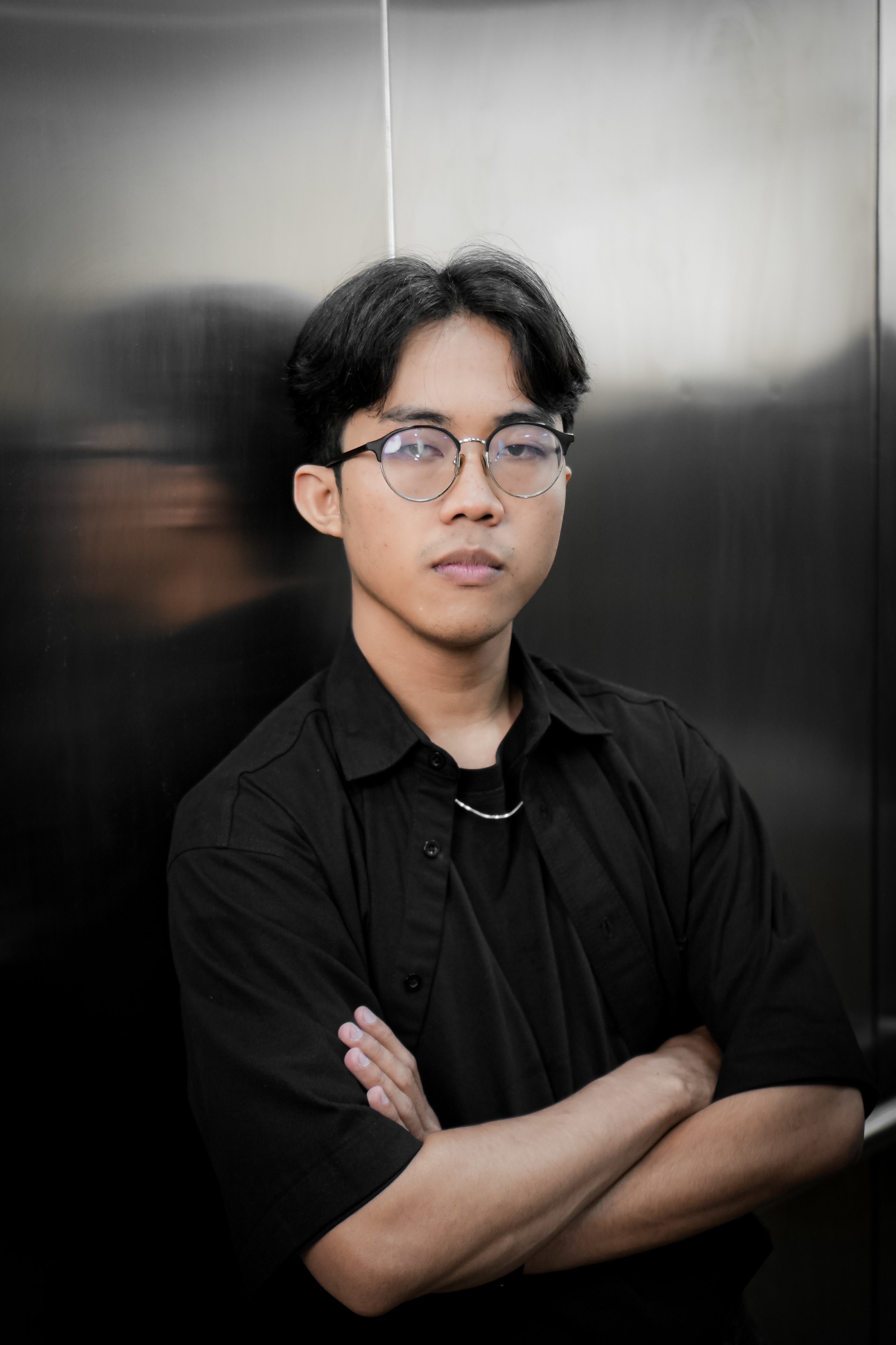 Young man with dark hair and glasses, wearing a black shirt, crossing his arms in front of a metallic elevator wall.