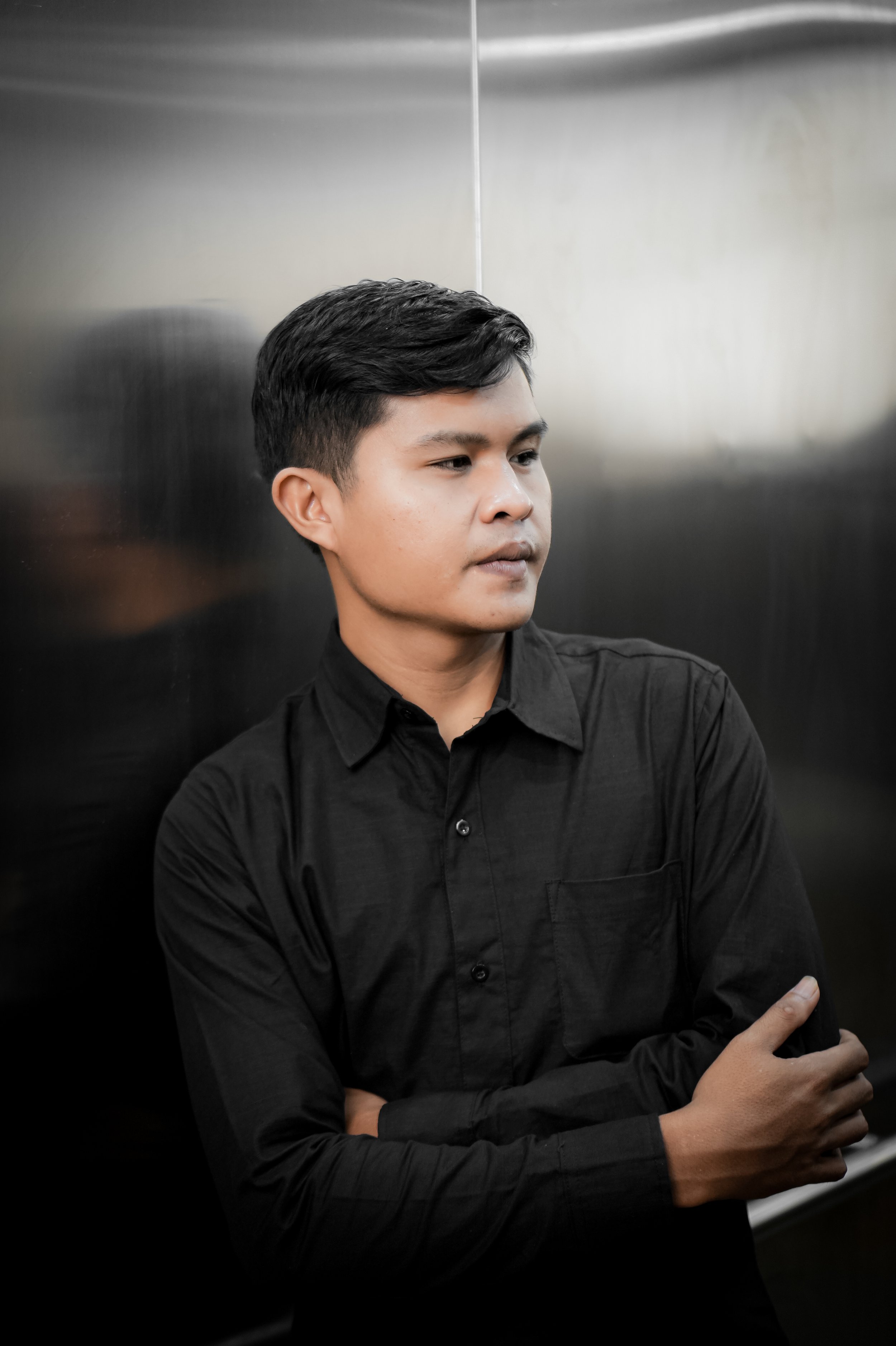 A young man with black hair wearing a black shirt, standing with arms crossed, in front of a metallic background.