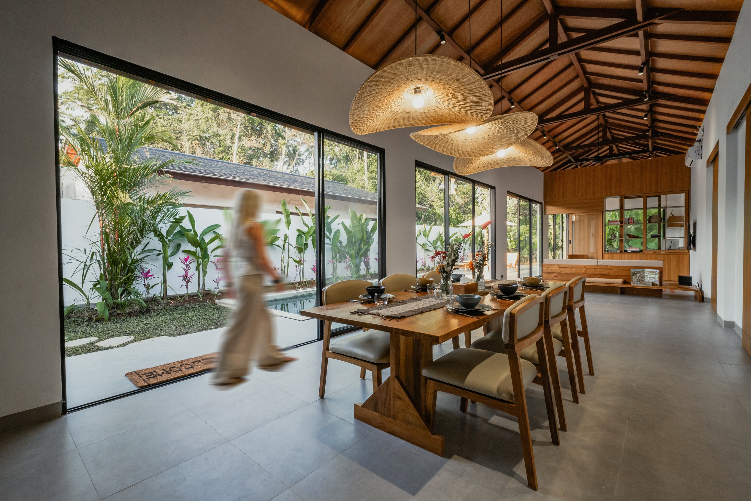 Modern dining room with large wood table, upholstered chairs, hanging woven lamps, and floor-to-ceiling windows overlooking a garden with plants.