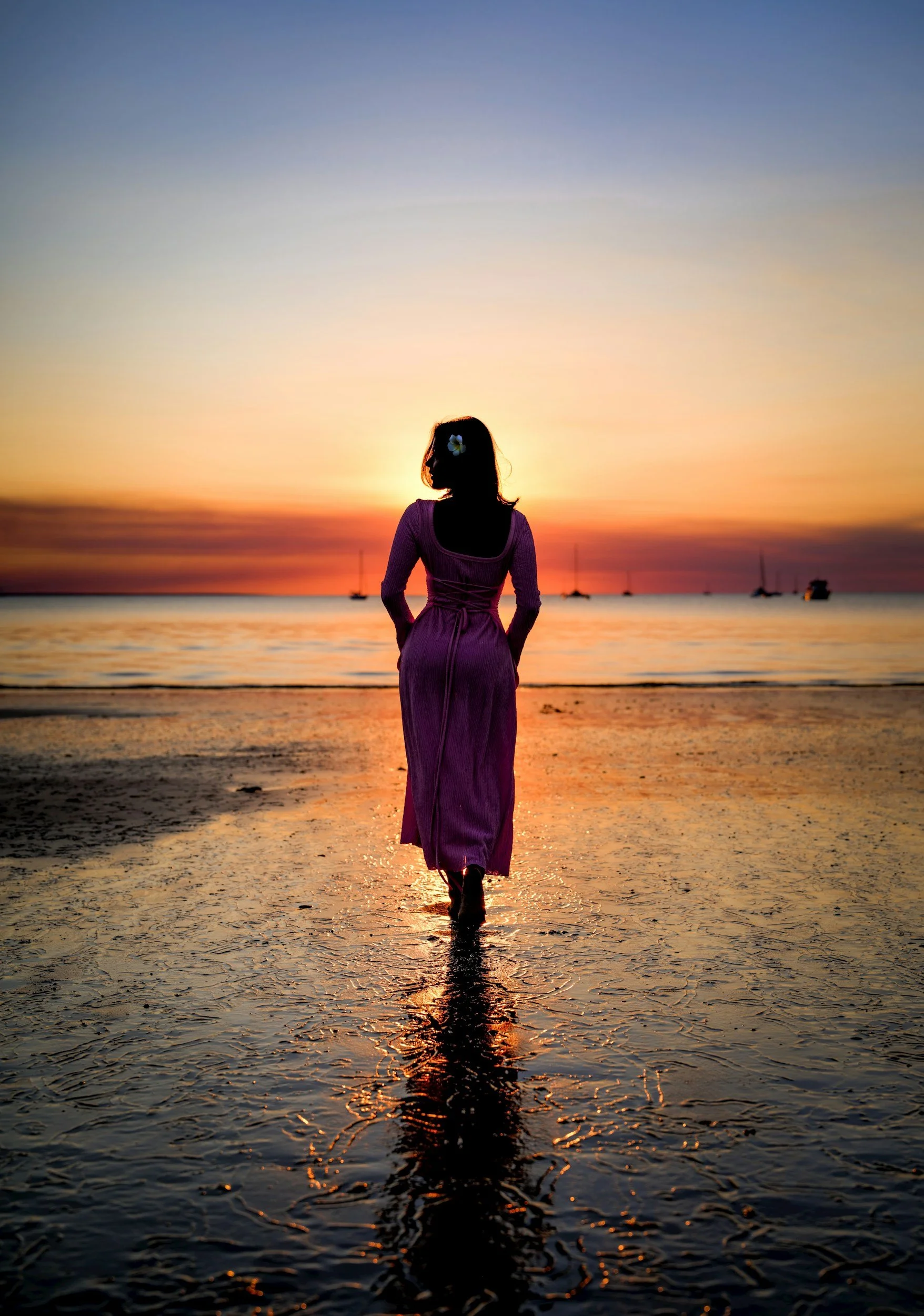 A woman in a purple dress walking on a beach at sunset, with sailboats in the distance and the sky colored in shades of orange, yellow, and blue.