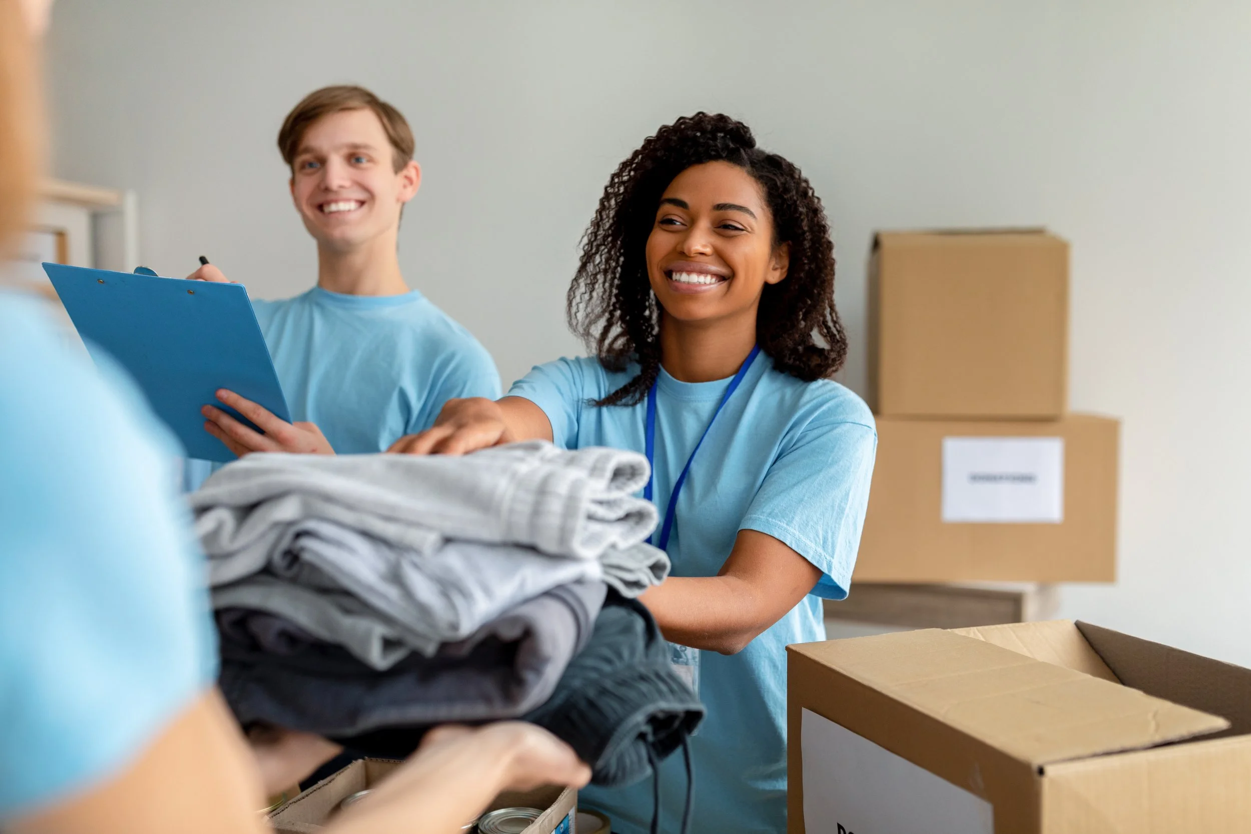 Two smiling volunteers dressed in blue shirts are packing clothes into boxes, with moving boxes stacked in the background.