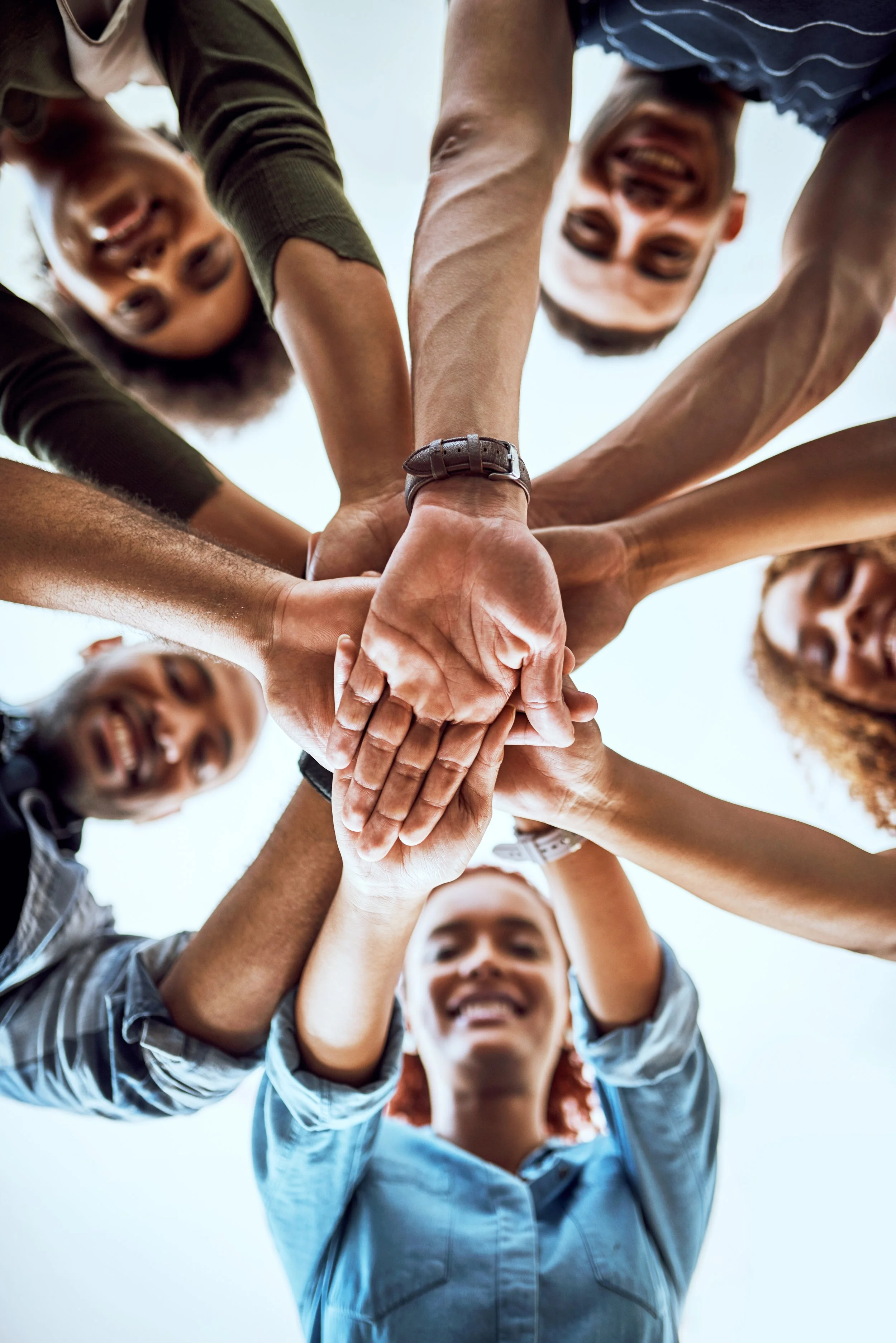 A diverse group of people in casual clothing forming a circle and stacking their hands in the middle, viewed from below against a clear sky.