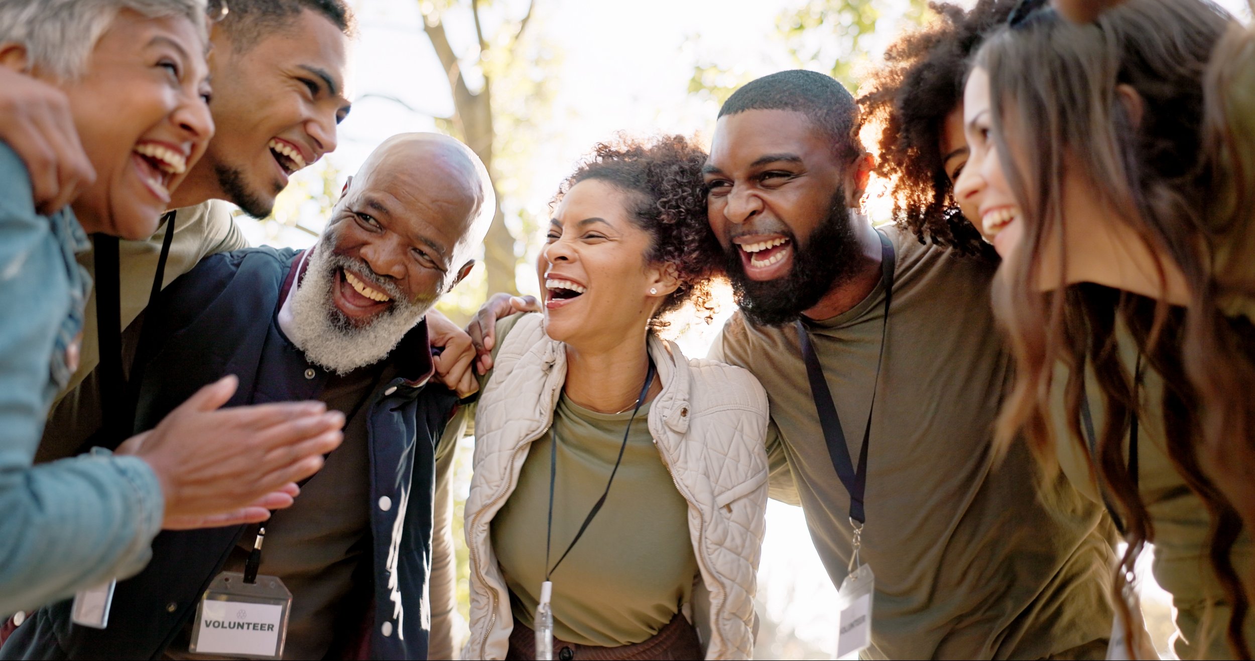Group of diverse people smiling and laughing closely together outdoors, some wearing volunteer badges.