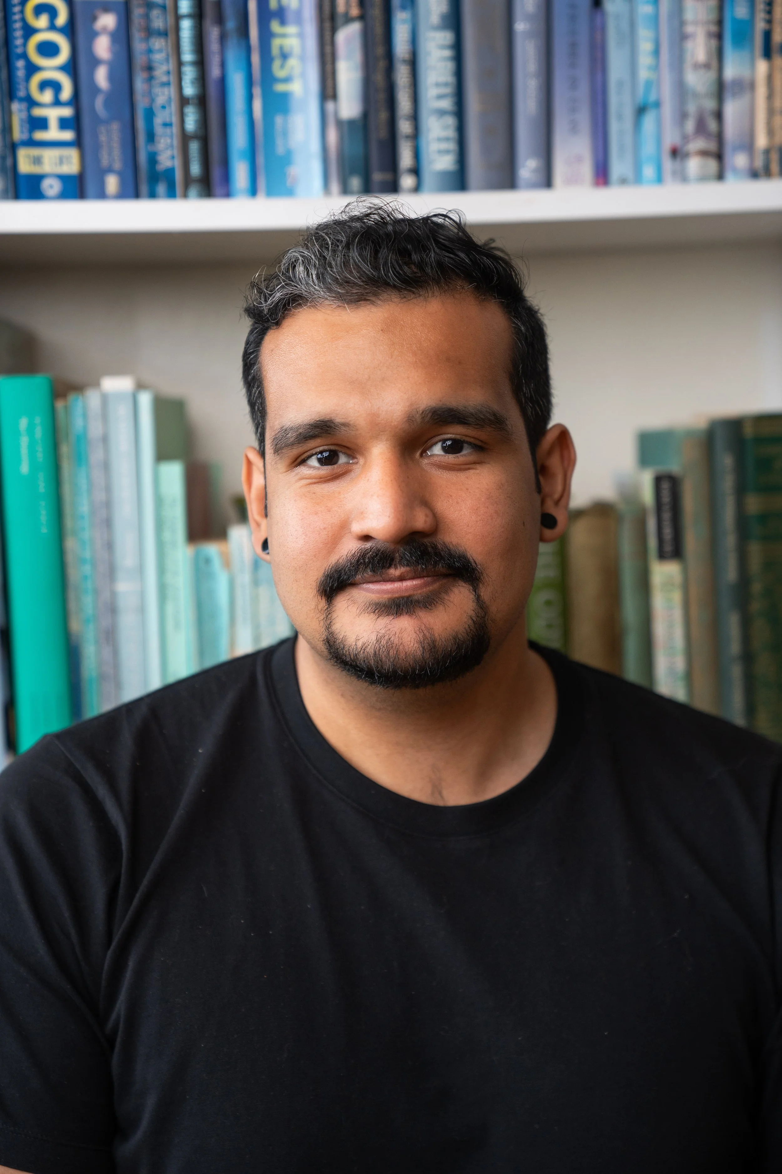 A portrait of a man with dark hair and a mustache, wearing a black shirt, with a bookshelf filled with books in the background.