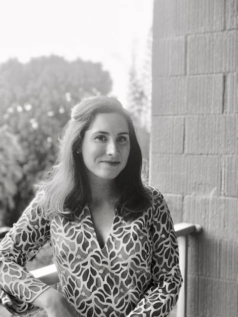 A woman with long hair wearing a animal print blouse, standing next to a brick wall outdoors.