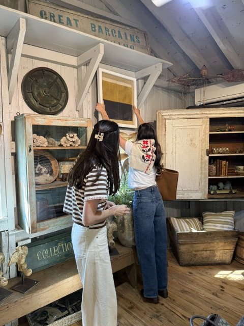 Two women hanging a framed artwork on a wall in a vintage store with shelves filled with decorative objects and books.