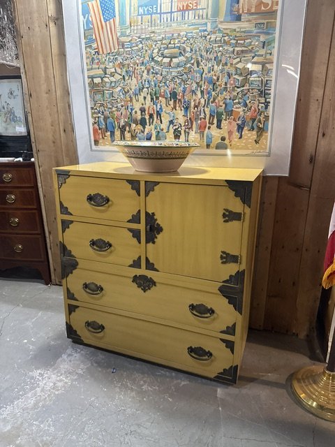 A yellow vintage dresser with black hardware and decorative accents, placed against a wooden wall inside a store. A bowl rests on top of the dresser, and an American flag is partially visible on the right. A framed picture of a busy street scene with crowds and signs, including NYSE, hangs above the dresser.