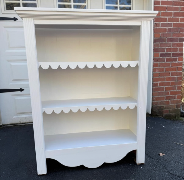 Empty white wooden bookshelf with three shelves, decorative scalloped edges, standing outdoors on driveway near garage and brick wall.