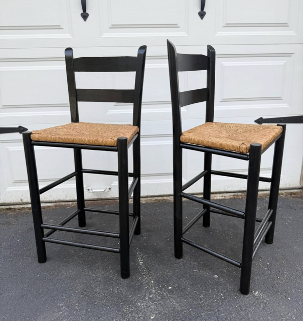 Two black wooden bar stools with woven straw seats are placed outdoors on a concrete surface in front of a white garage door.